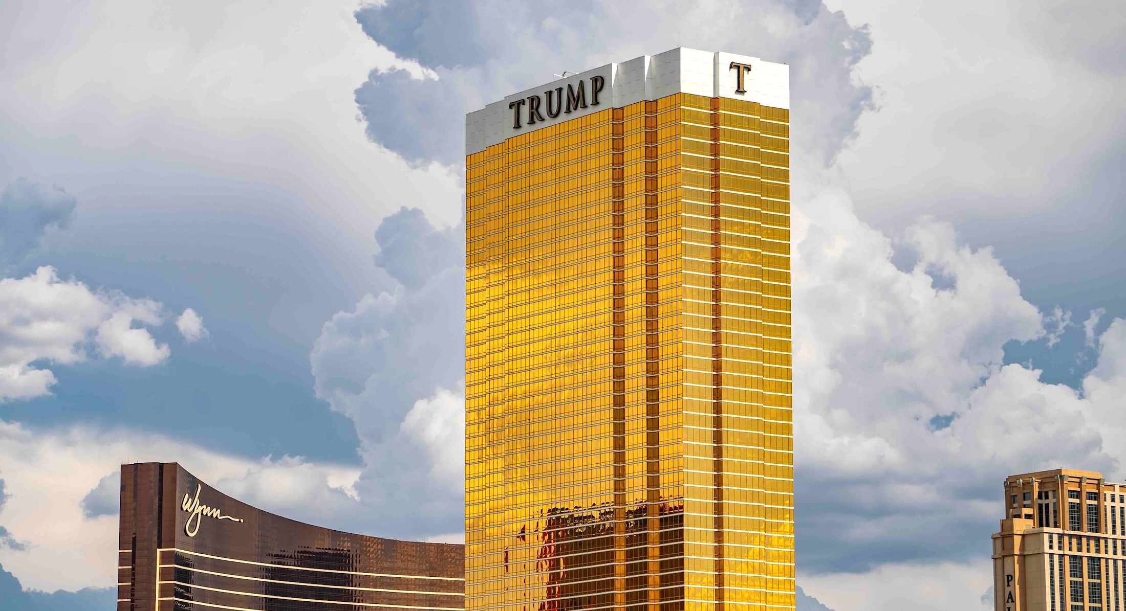 A tall gold tower with the word &lsquo;Trump&rsquo; at the top in black against a white background stands against a blue sky with white clouds.