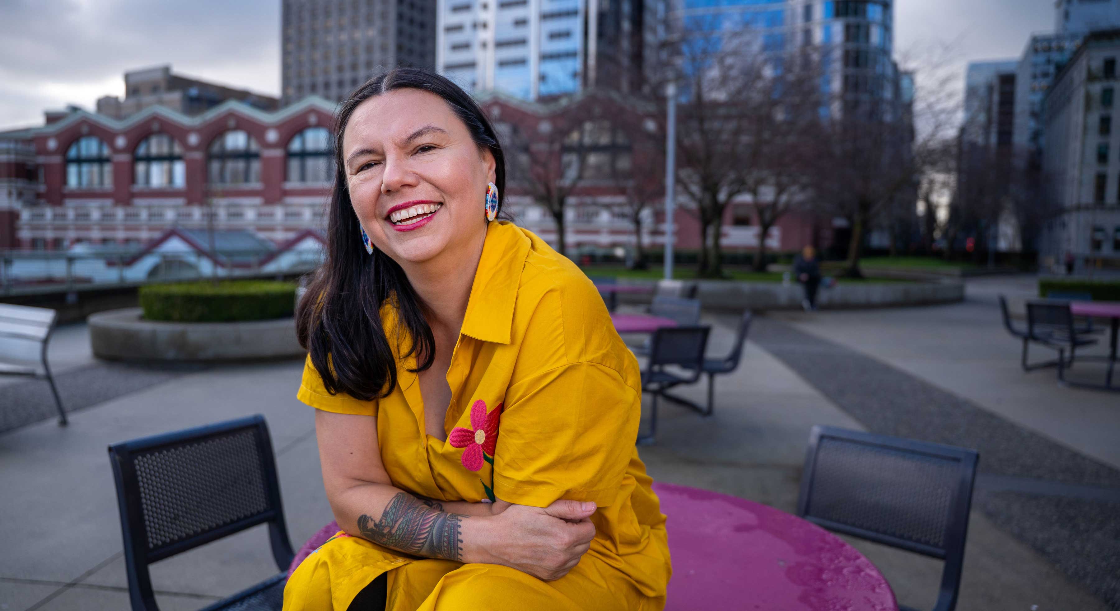 Eden Fineday is laughing, smiling widely. She has long dark hair and is wearing a yellow dress with floral embroidery. She is seated on a magenta table in an outdoor plaza with her arms folded in her lap.