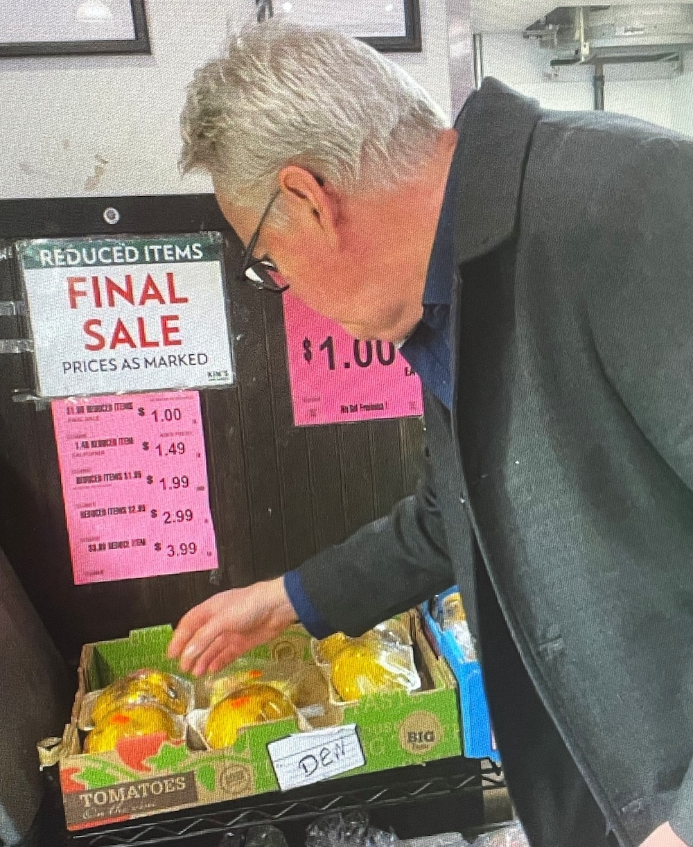 Steve Burgess is wearing a black jacket and has his back turned to the camera as he inspects a discount box of fruit at a grocery store. 
