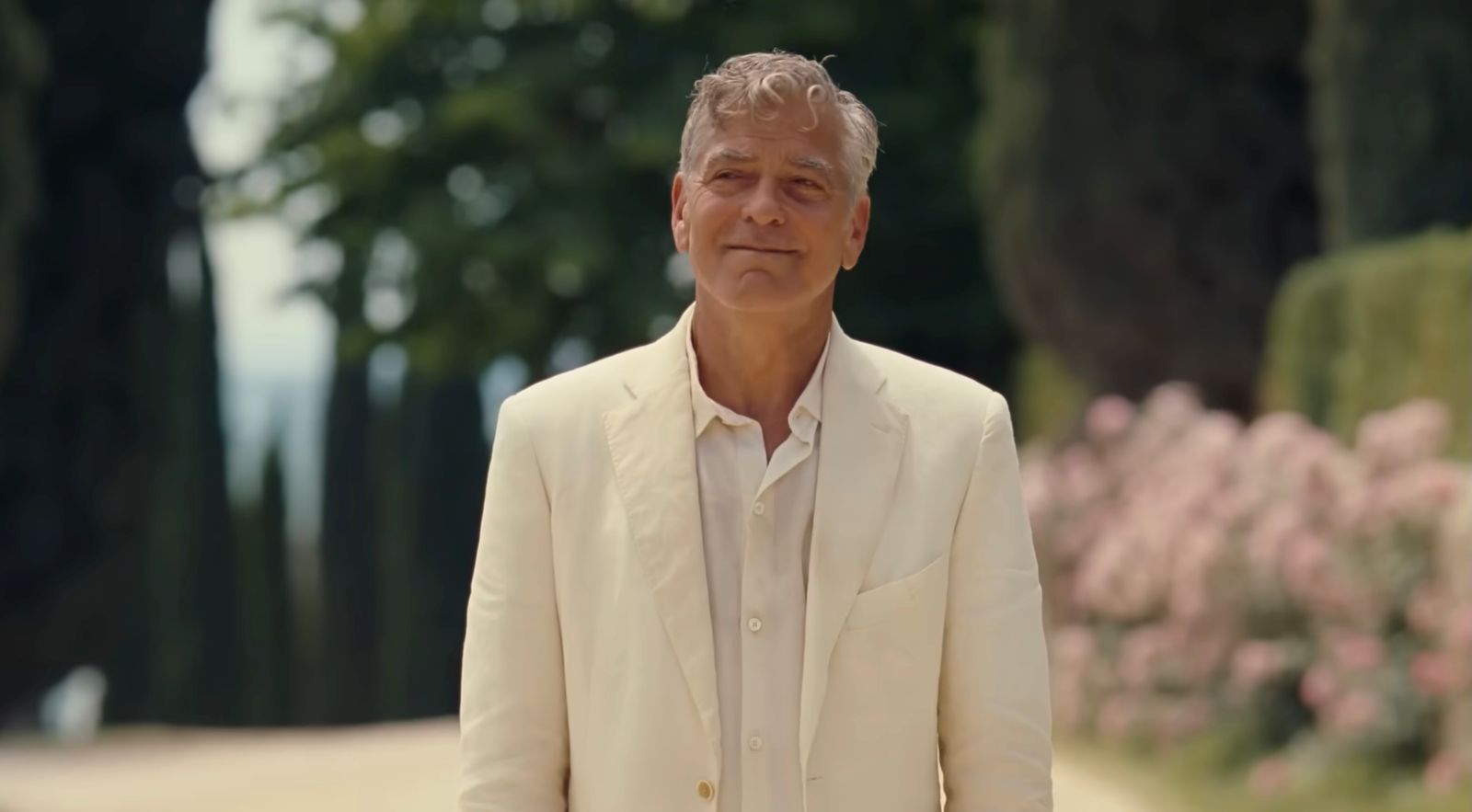 George Clooney is wearing a cream-coloured suit. He has white curly hair and tanned skin. He is standing outdoors with the background in soft focus.