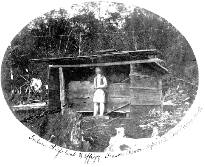 A rectangular wooden grave house structure with a carved figure in front.