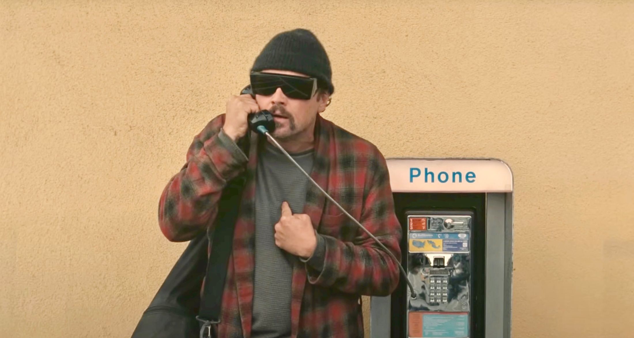 Actor Leonardo DiCaprio is wearing wide sunglasses, a toque and a red flannel shirt over a grey T-shirt. He is pointing at his chest while speaking into a payphone against a light yellow textured wall.