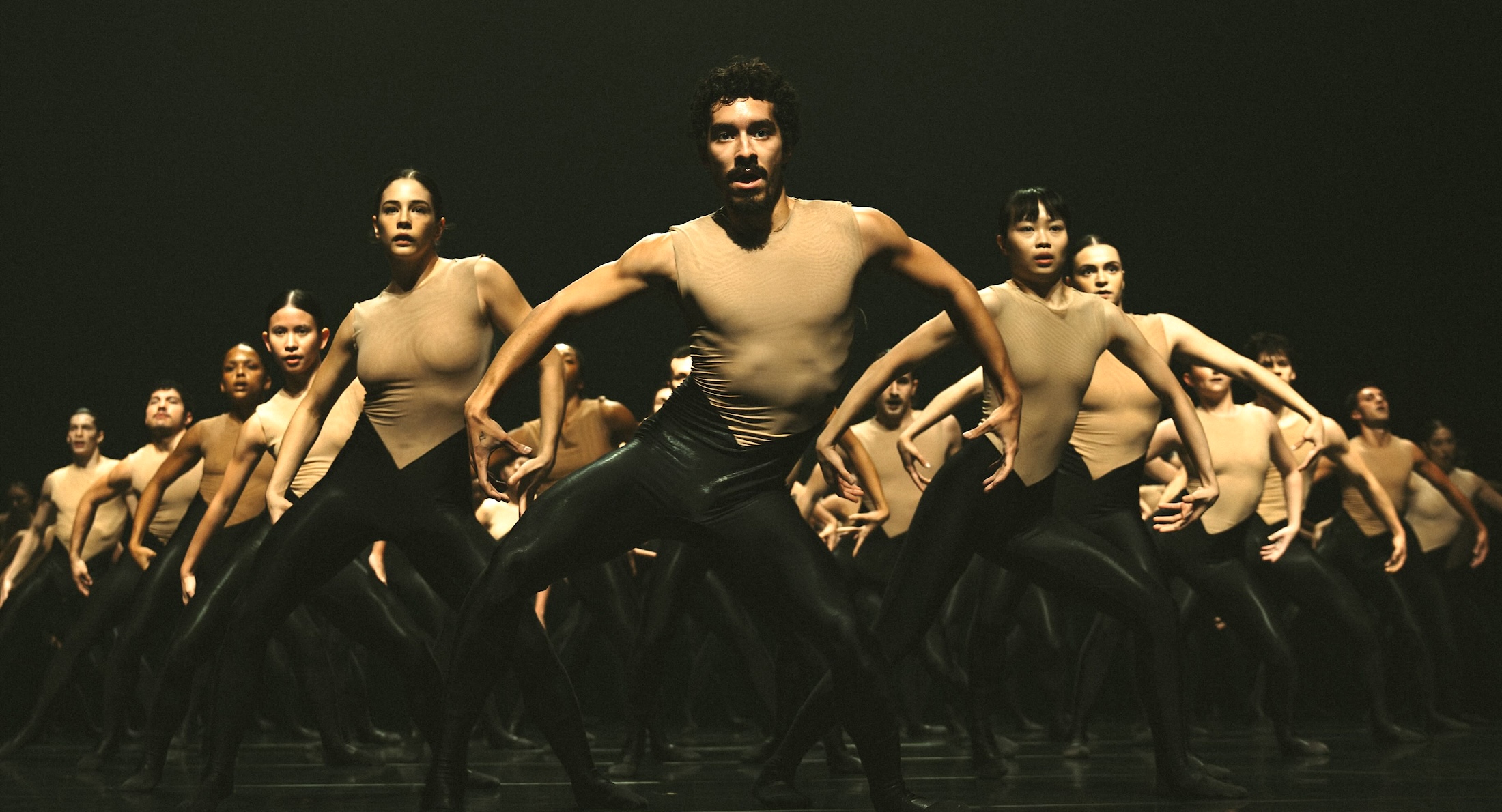 A troupe of dancers in matching beige tops and black pants stand in formation with their arms out to their sides against a black background.