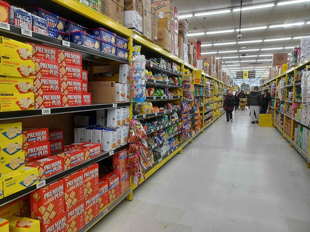 The aisle of a discount grocery store with yellow signage features rows of crackers in yellow and red packages to the left of the frame.