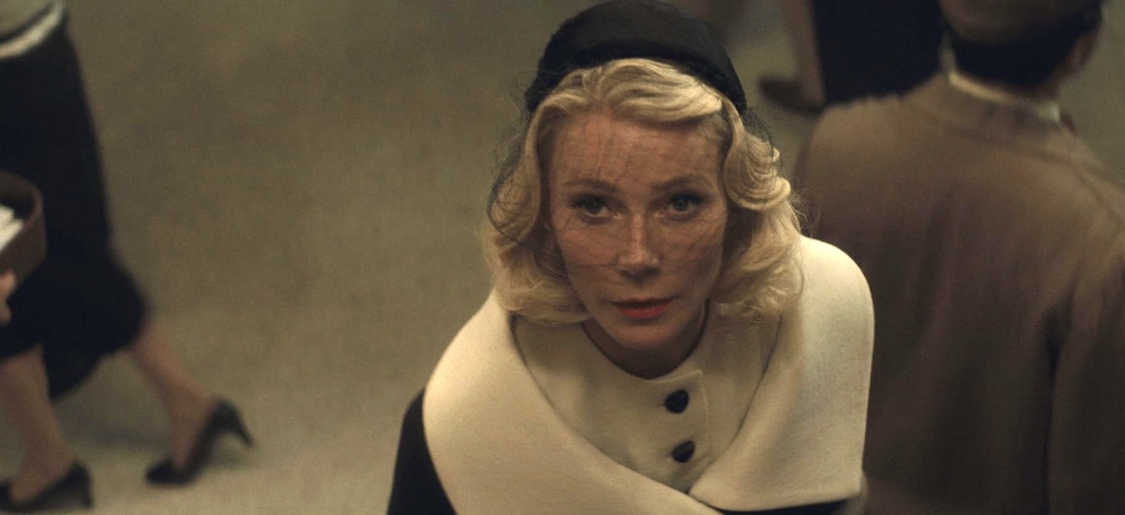Actor Gwyneth Paltrow is dressed in a 1950s-style car coat with a wide cream collar over a cream-coloured shirt with large buttons. She is wearing a black hat with a black veil. She has a light skin tone and short curly blond hair.
