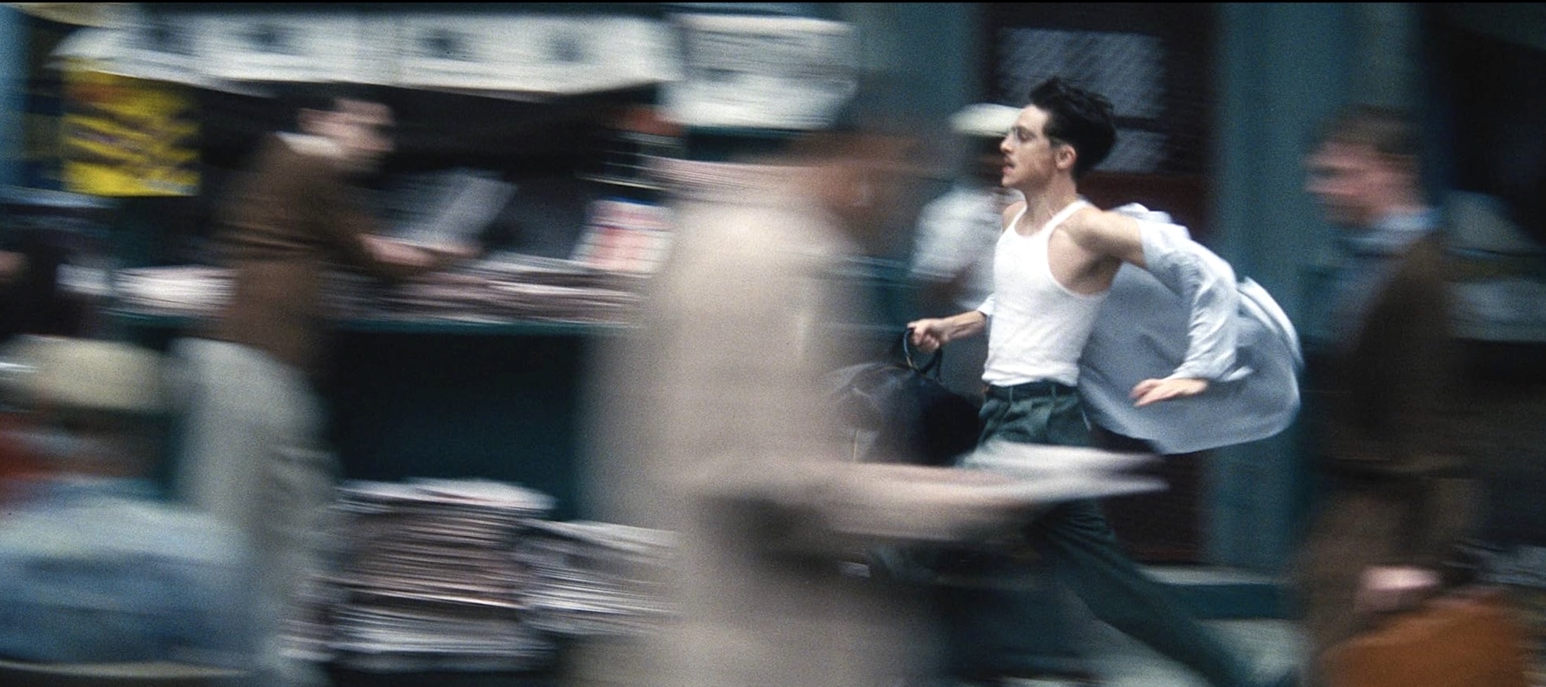 Actor Timoth&eacute;e Chalamet runs towards the left of the frame and is shot from the side. He has short dark hair, glasses and a moustache. He is wearing a white undershirt under a light blue button-down shirt and holding a bag while he runs down a crowded city street; the people and landmarks around him are blurry.