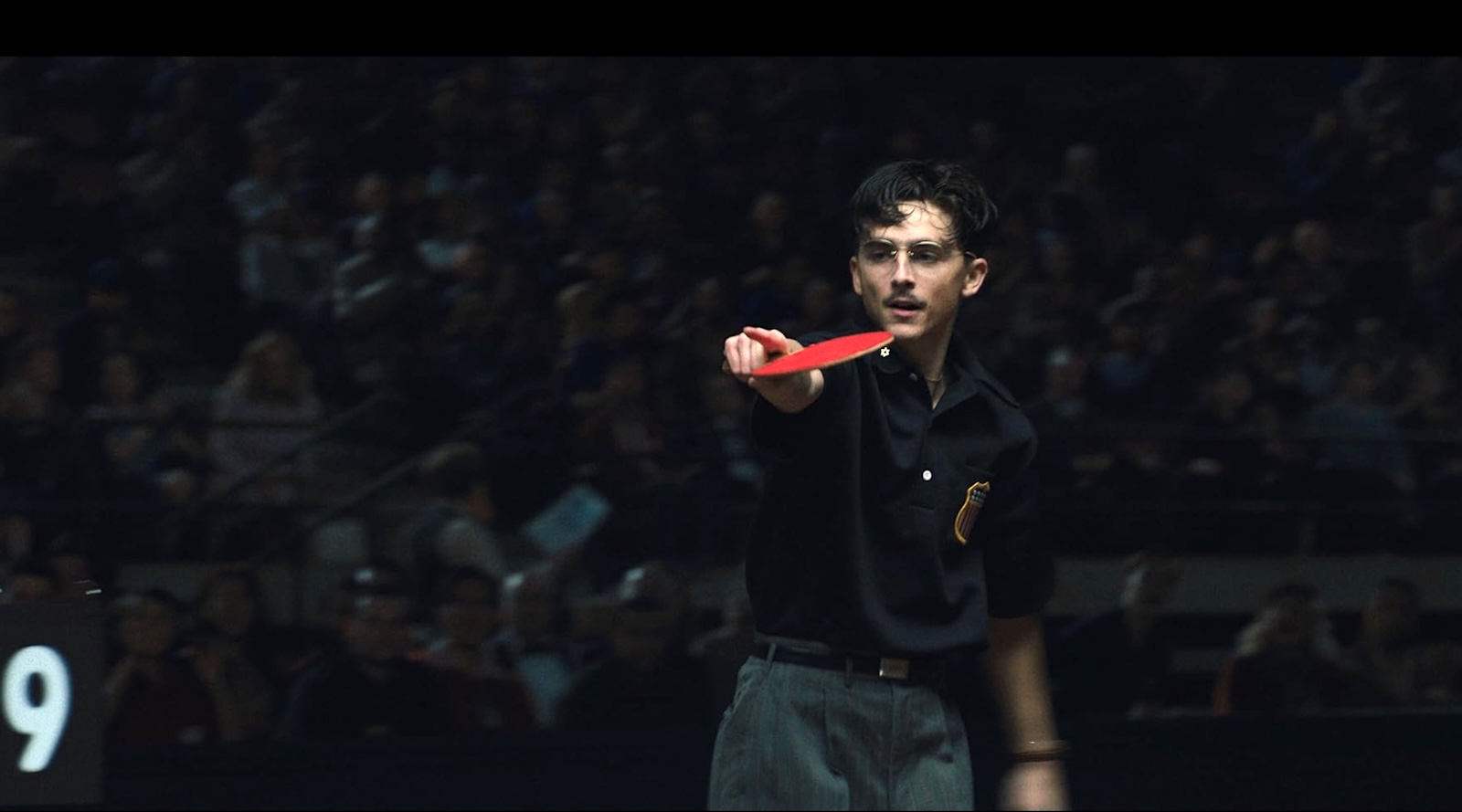 Actor Timoth&eacute;e Chalamet stands holding a red ping-pong paddle in a black short-sleeved shirt against a dark background. He has short dark curly hair and glasses and is pointing forward with his racket.