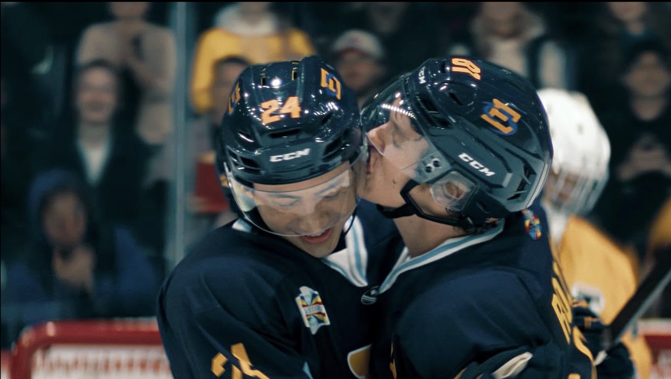 Two actors in dark blue hockey uniforms are on the rink together. Connor Storrie, right, kisses Hudson Williams, left, on the cheek.
