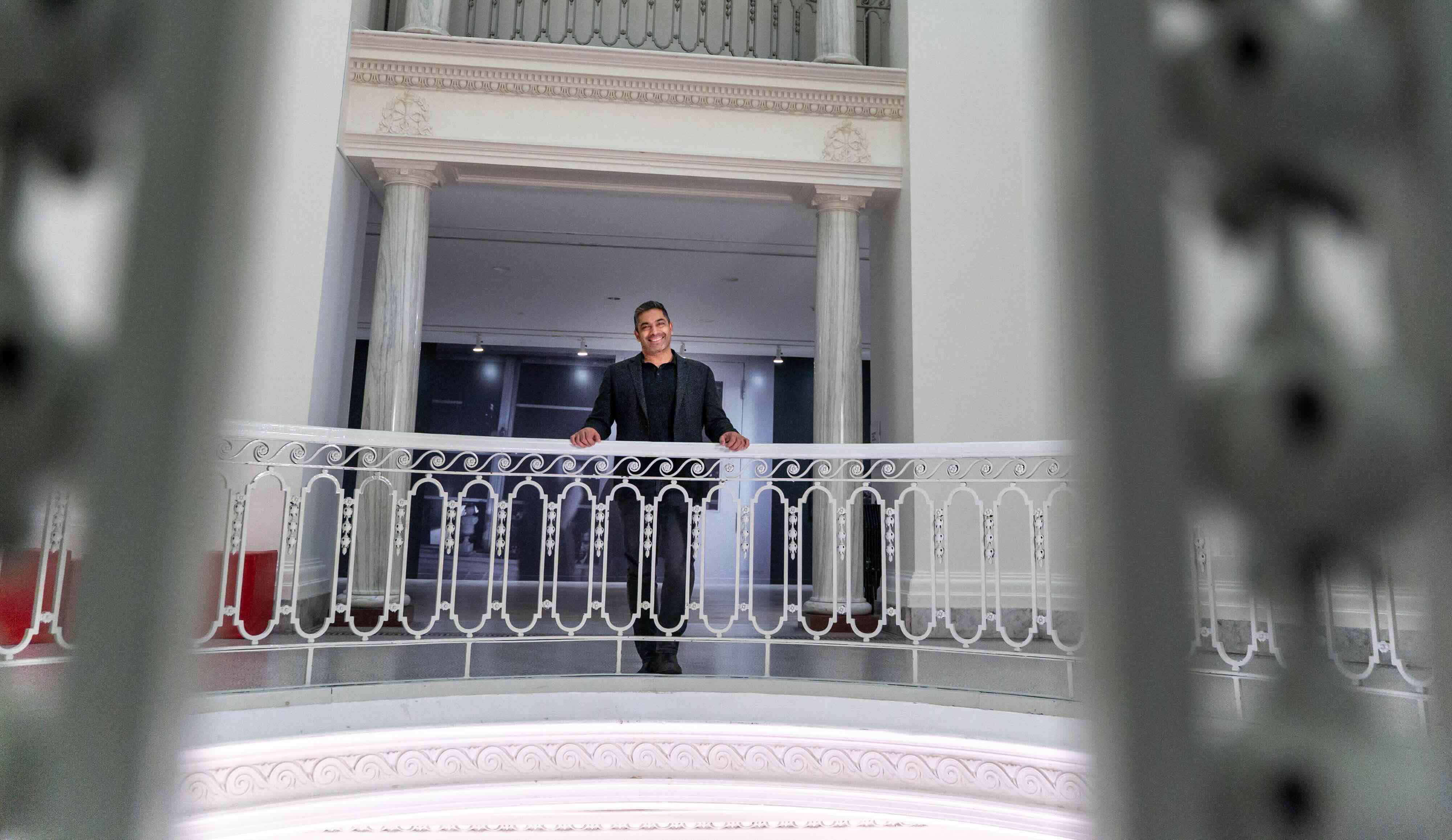 Sirish Rao is standing against an ornate white balcony railing in the midlle of a tall white rotunda at the Vancouver Art Gallery. He is wearing a dark suit and smiling.