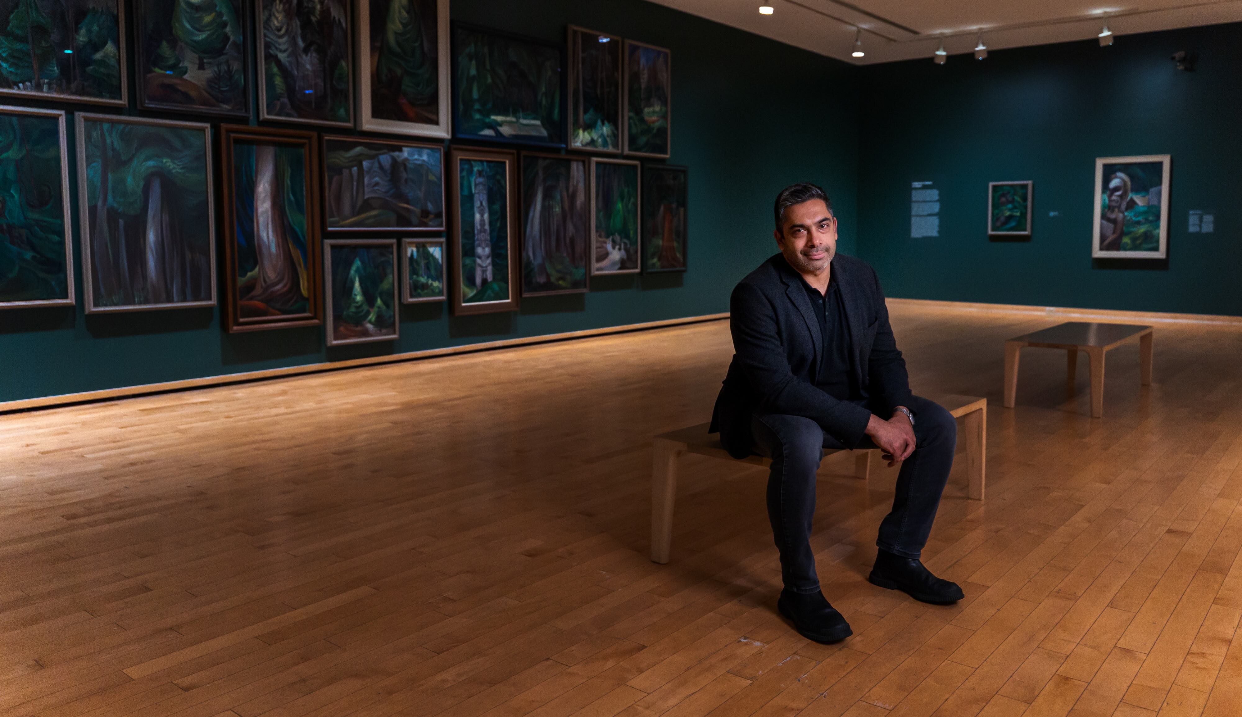 Sirish Rao is seated on a bench in a gallery space with wood floors and dark teal walls on which framed oil paintings of trees are hung.