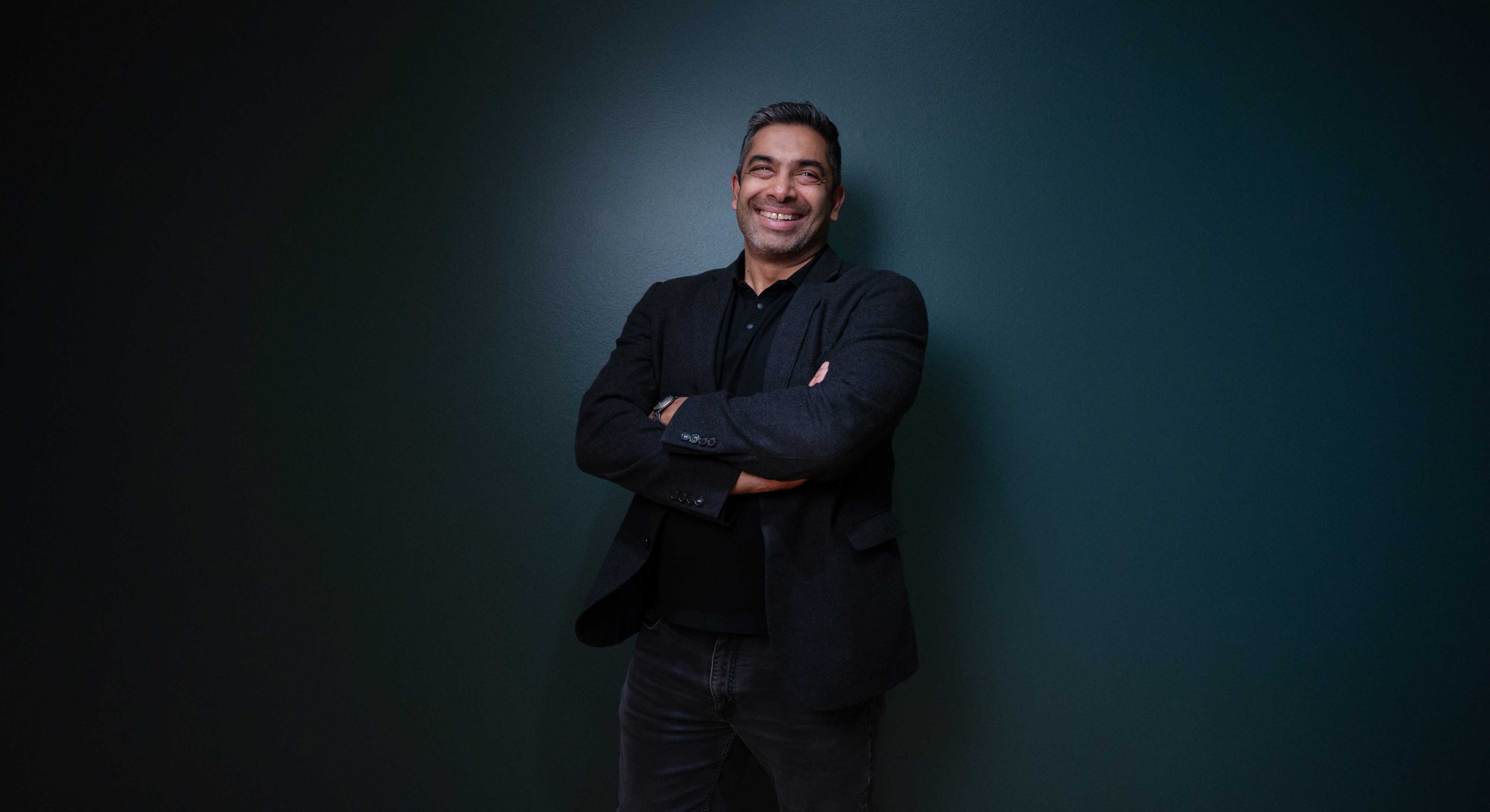 Sirish Rao has short dark hair. He stands against a dark teal wall with his arms folded, smiling.