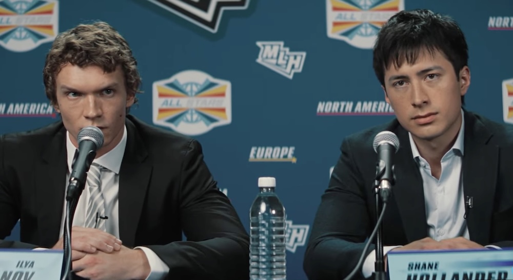Two actors are wearing dark suits over light shirts. They are seated at a table behind microphones at a hockey press conference. On the left, Connor Storrie is speaking into a microphone. He has blond curly hair. On the right, Hudson Williams is listening. He has short dark hair.
