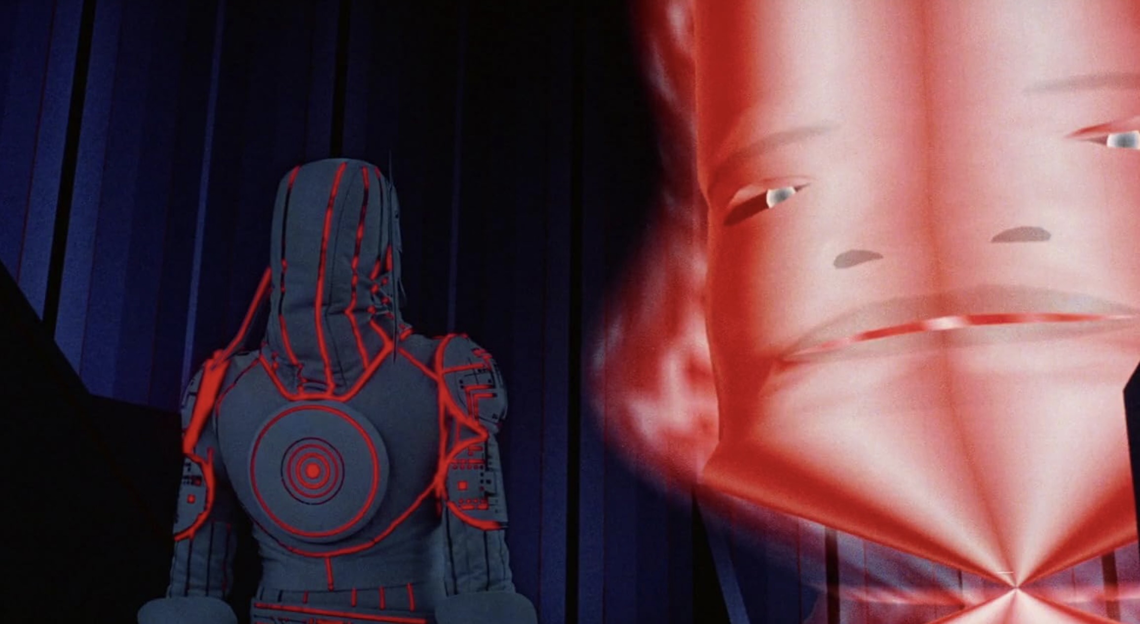 A man in a tight grey costume with black-lit red details stands in a darkened room against which a large red digital robot face is projected.