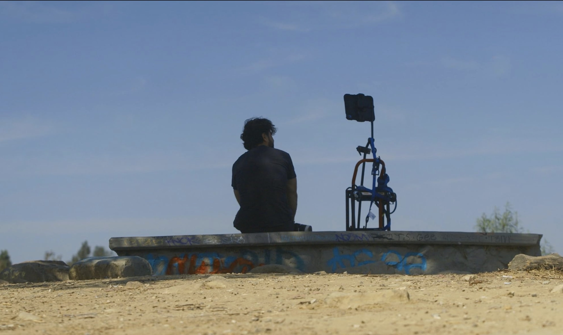 A man with curly dark hair sits with his back to the camera at the left of the frame. He looks towards the mounted digital device next to him. They are seated together on a concrete structure against a blue sky. 