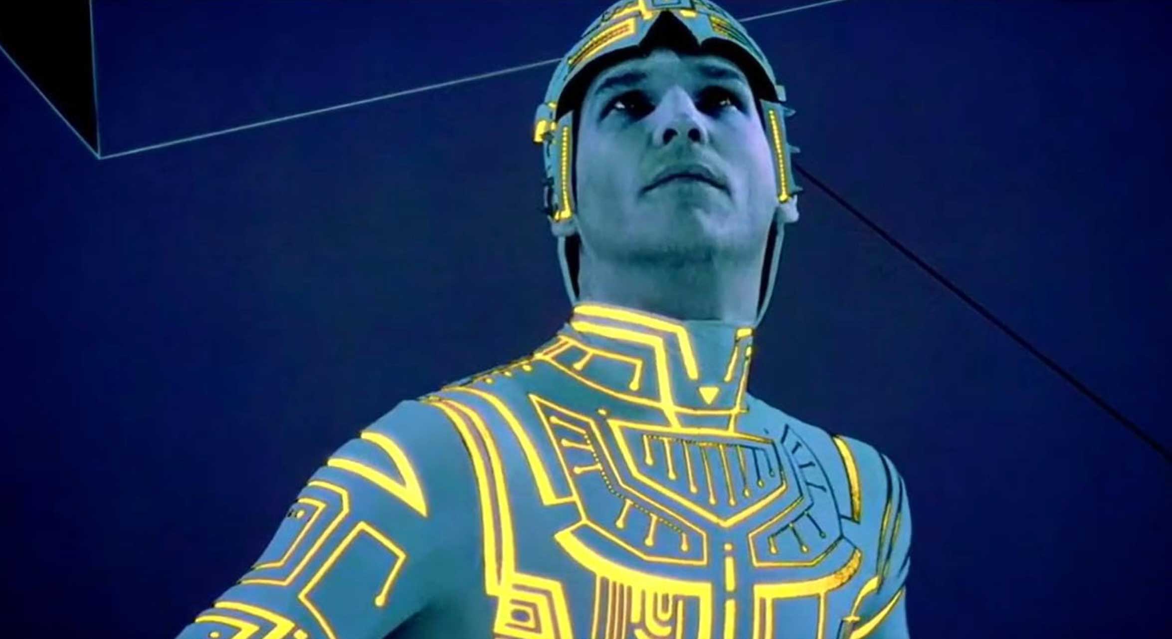 Actor Jeff Bridges is wearing a tight light blue costume and matching headpiece with a glowing black lit yellow pattern. He is looking up and standing against a dark blue background.