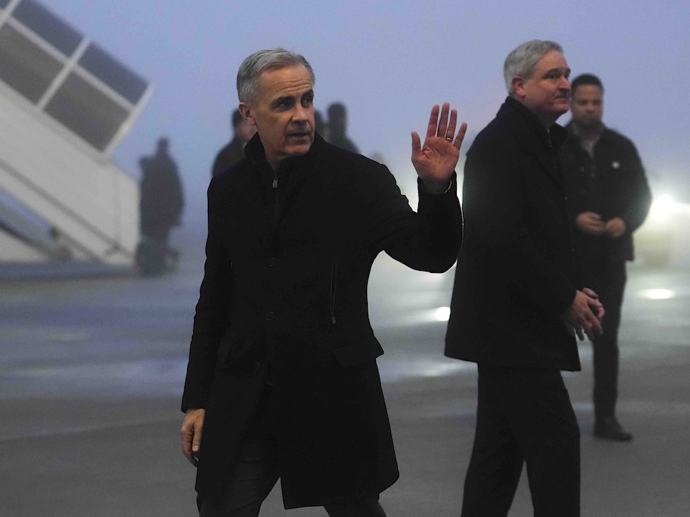 Mark Carney is waving as he walks along an airport tarmac. He is wearing a black jacket and trousers and the sky is dim.