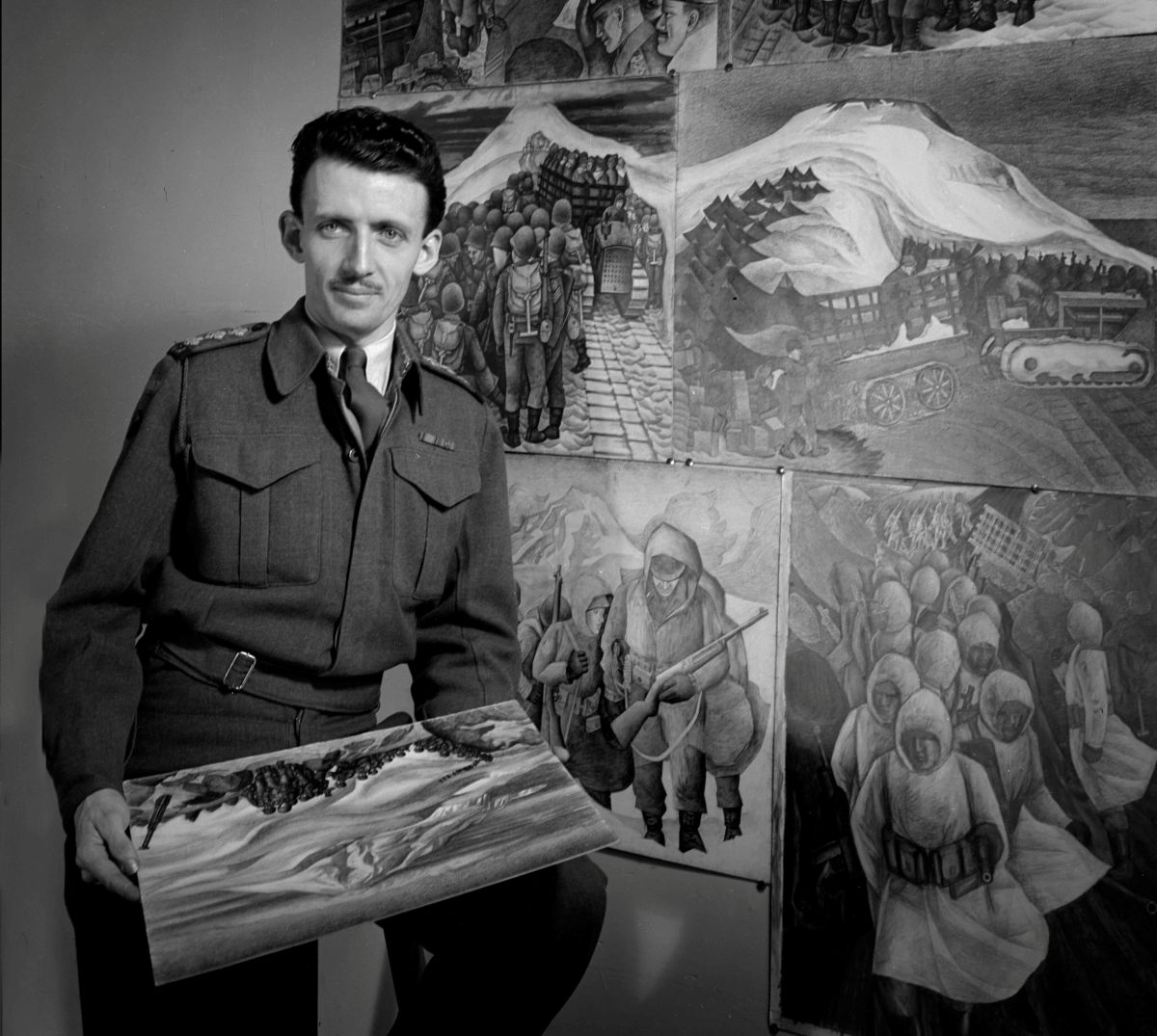 E.J. Hughes stands next to a wall featuring his paintings depicting the events of the Second World War. He is standing in a military suit, holding one of his landscape paintings.
