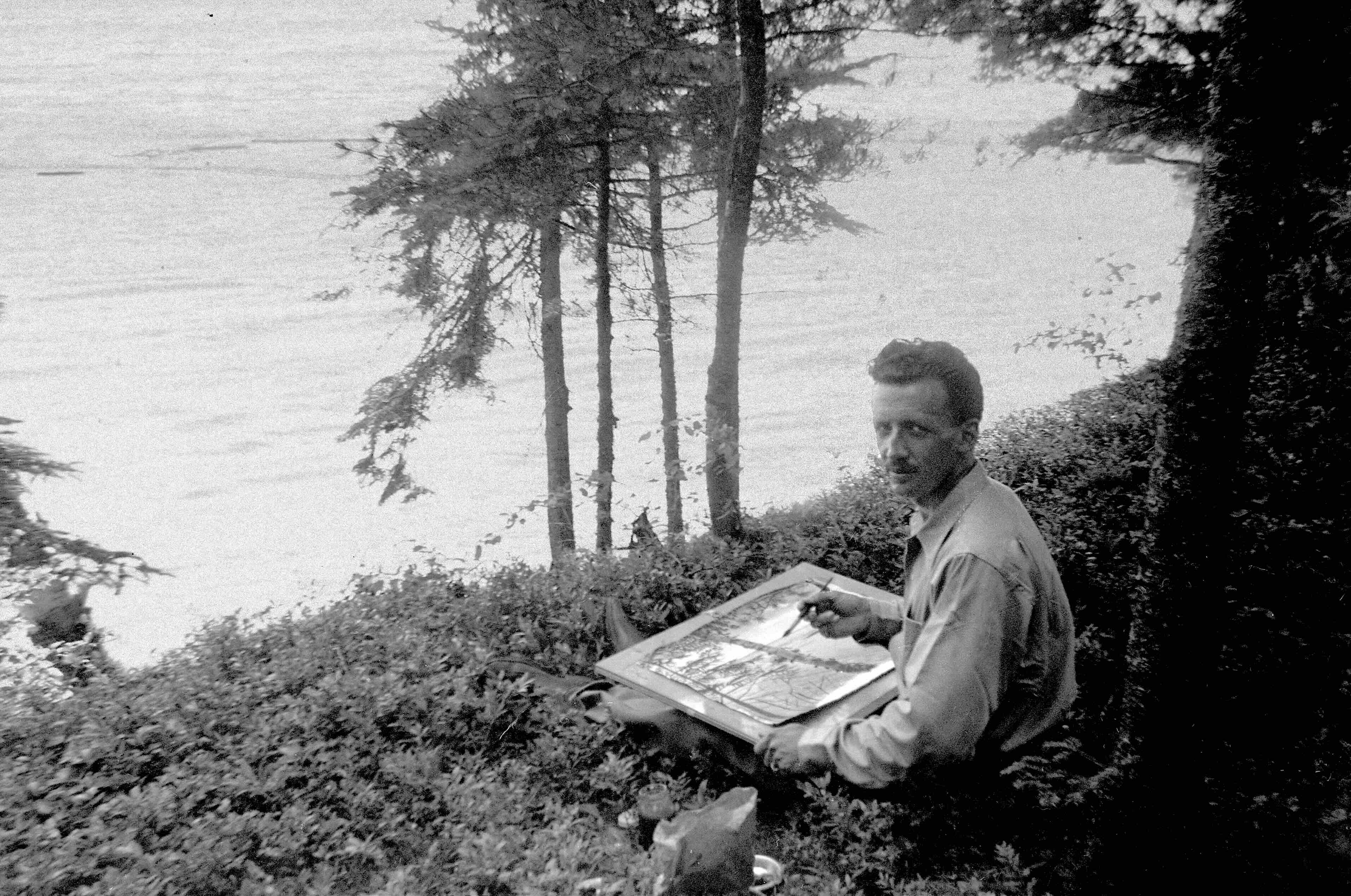 E.J. Hughes has short dark wavy hair and a moustache. He is seated outdoors overlooking a body of water, painting. He is turning around to face the camera.