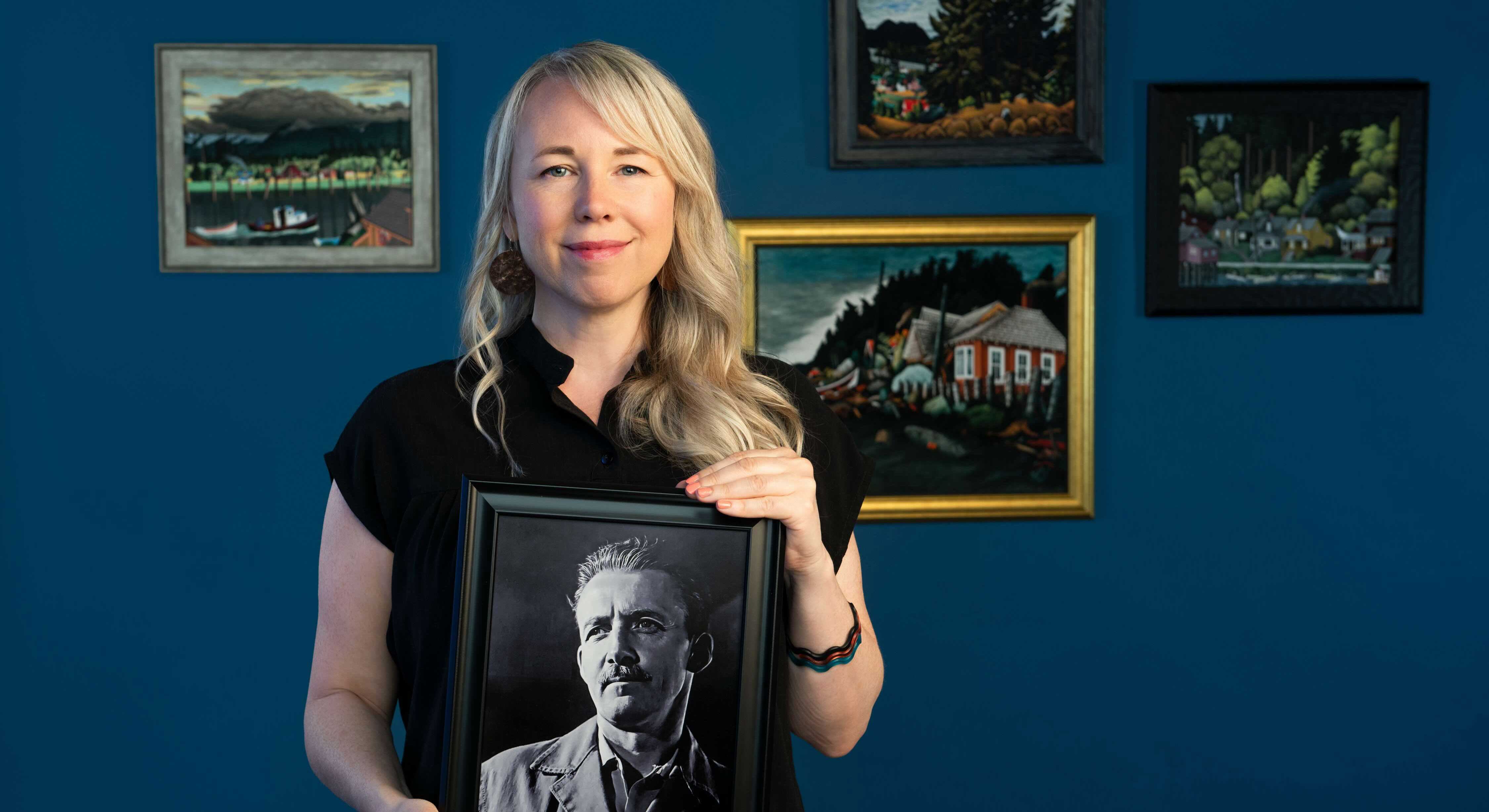 Jenn Strom has long blond wavy hair. She is smiling at the camera and holding a black-and-white portrait of E.J. Hughes, a man with short dark wavy hair and a moustache, wearing a suit. She stands against a dark blue wall hung with several small framed landscape paintings.