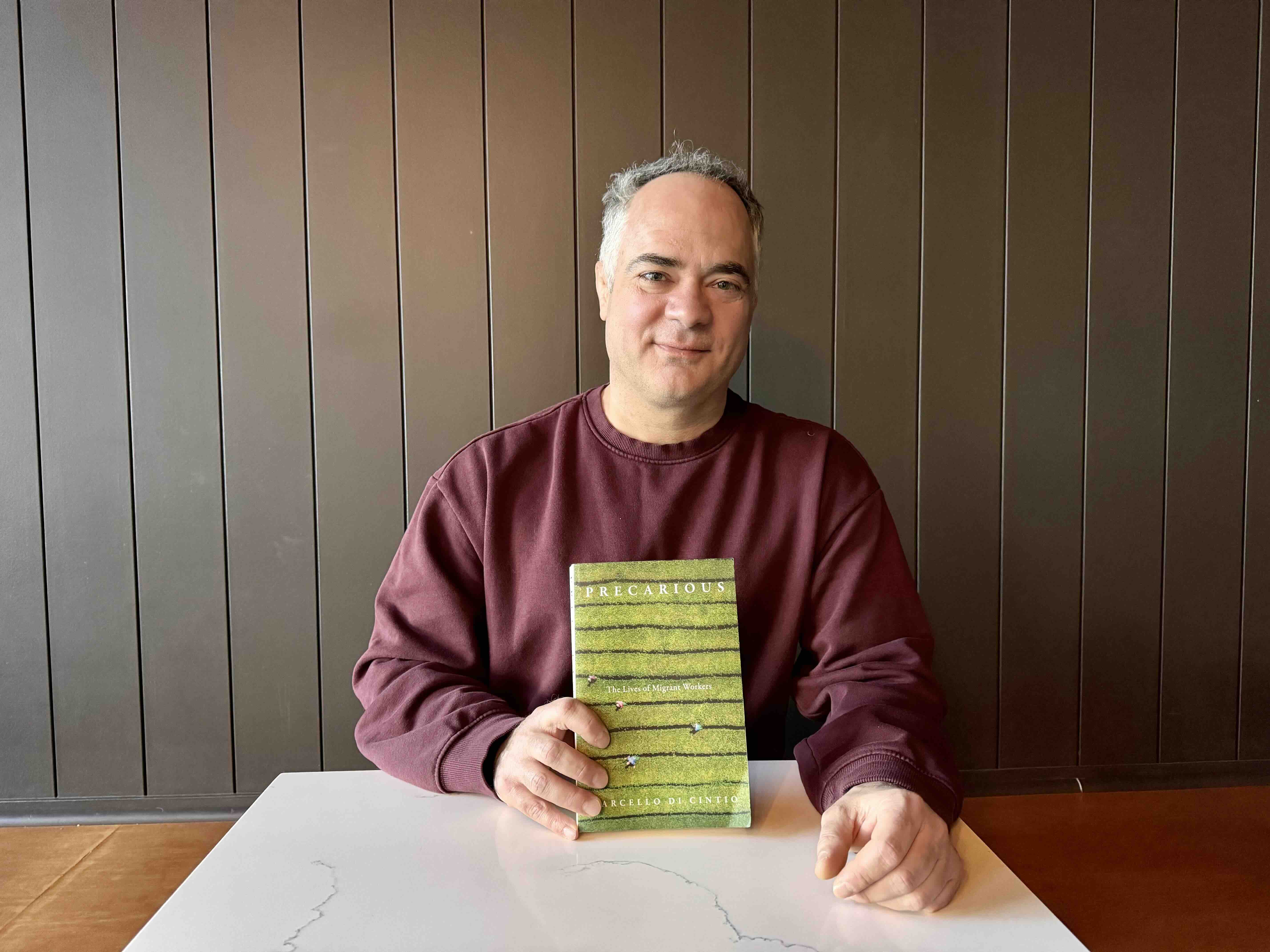 Marcello Di Cintio is seated at a table against a dark brown all. He has short grey hair and he is wearing a red crewneck cotton shirt. He is holding a copy of his book, Precarious: The Lives of Migrant Workers. It features an aerial photo of people working in a green field.