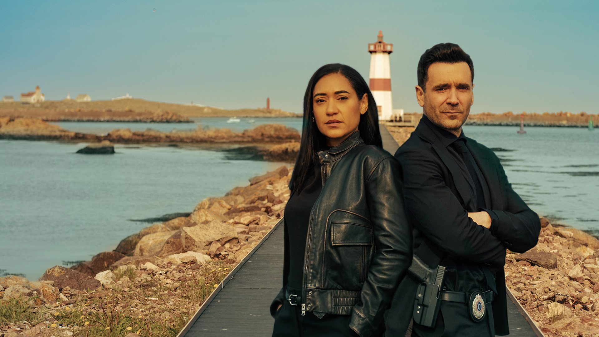 Actors Jos&eacute;phine Jobert, at left, and Allan Hawco, at right, stand together on a walkway to a lighthouse in the background. They are both dressed in back and looking at the camera with serious expressions.