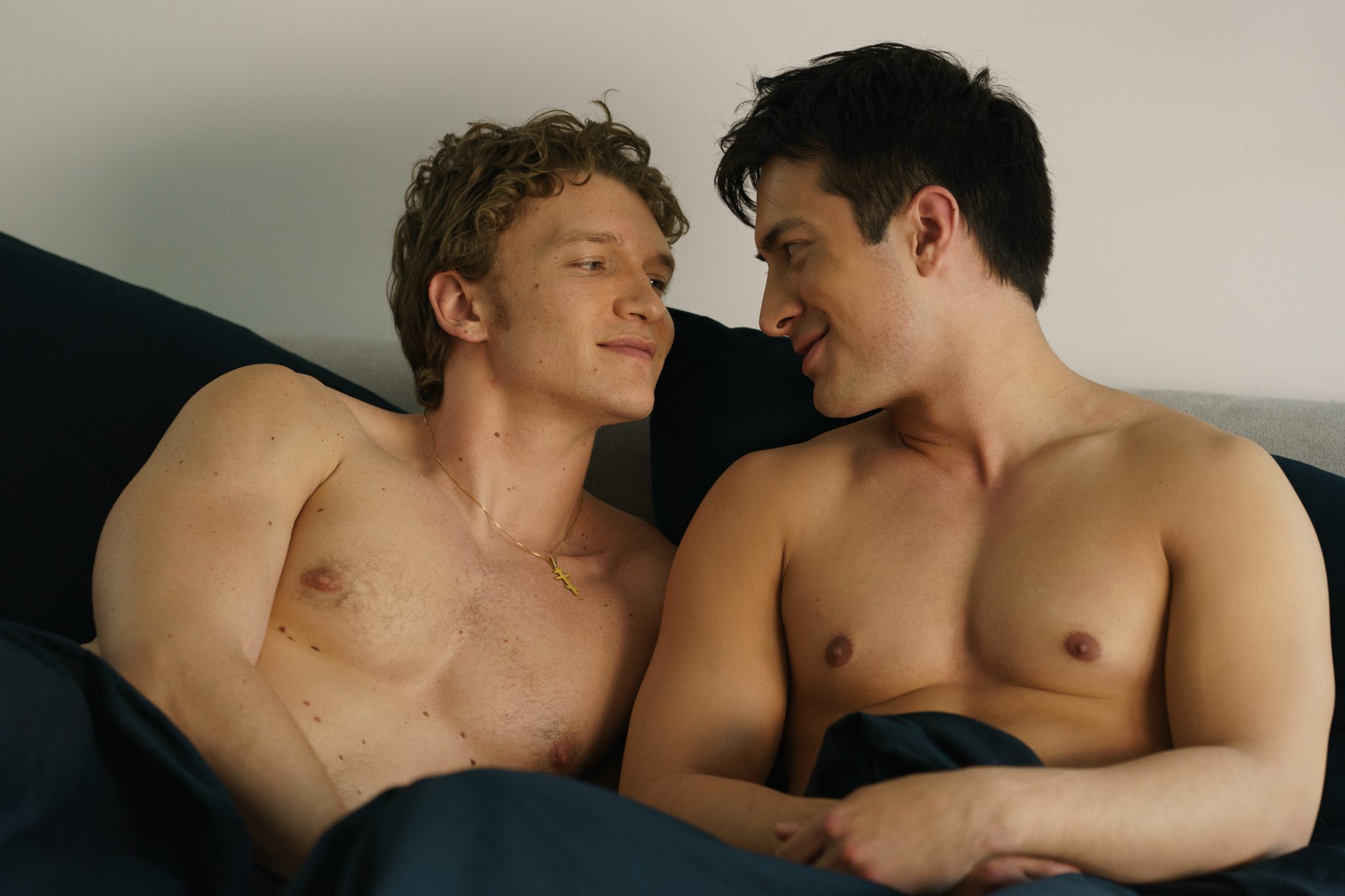 Actors Connor Storrie, at left, and Hudson Williams, at right, are seated together in bed with navy sheets.