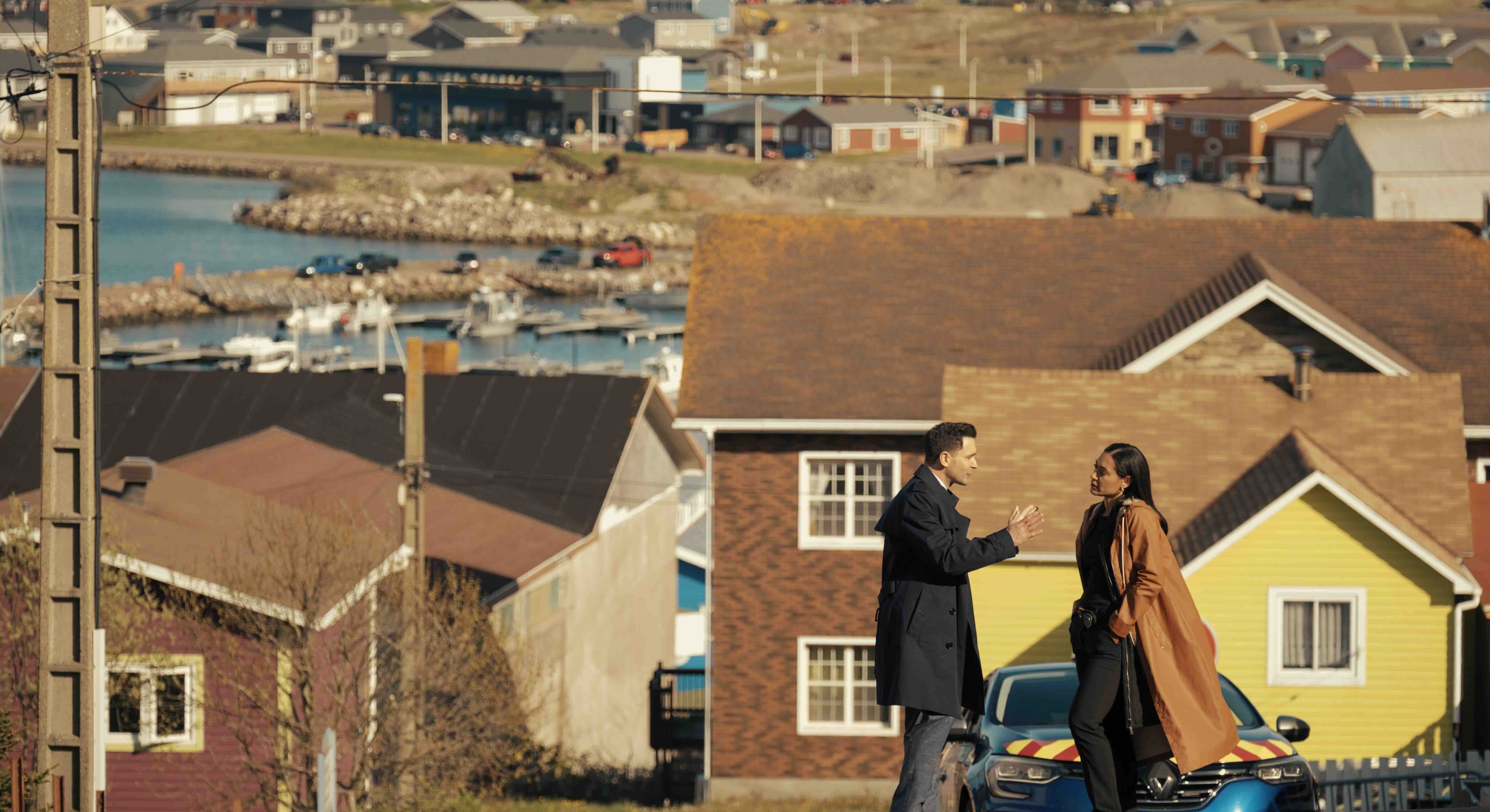 Actors Allan Hawco, at left, and Jos&eacute;phine Jobert, in conversation at the top of a hill on a residential street on the island of Saint-Pierre. Jobert has long dark hair and is wearing a long camel-coloured coat and Hawco is wearing a long black coat. Behind them is a yellow house and several other homes in a coastal community.