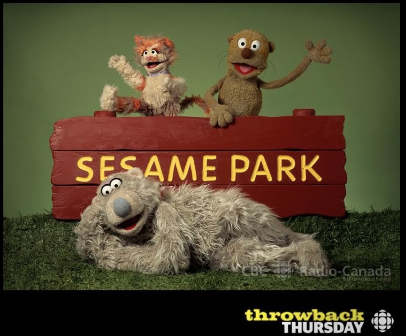 A cat and a rodent puppet wave from behind a red wooden sign that reads
&ldquo;Sesame Park&rdquo; while a polar bear puppet lies across the front.