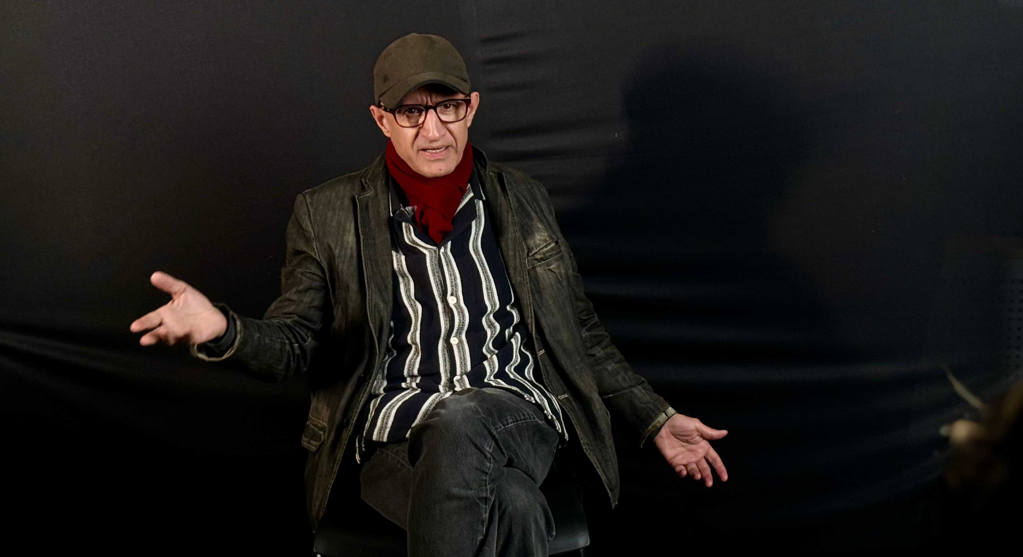 Hossein Martin Fazeli is seated against a dark background, mid-speech. He is wearing an olive-coloured ball cap, a red scarf and a black leather jacket over a black and white striped shirt. He has glasses and a medium skin tone.