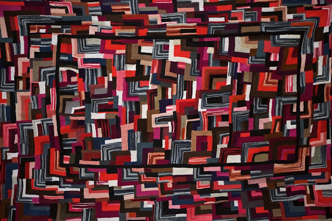 A large-format piece of abstract embroidery work features bold red, pink and black tones and dense geometrical patterns.