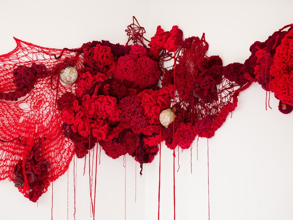 A piece of red textile artwork against a white background uses dark and brighter shades of red crocheted wool, velvet, ceramics and fabric.
