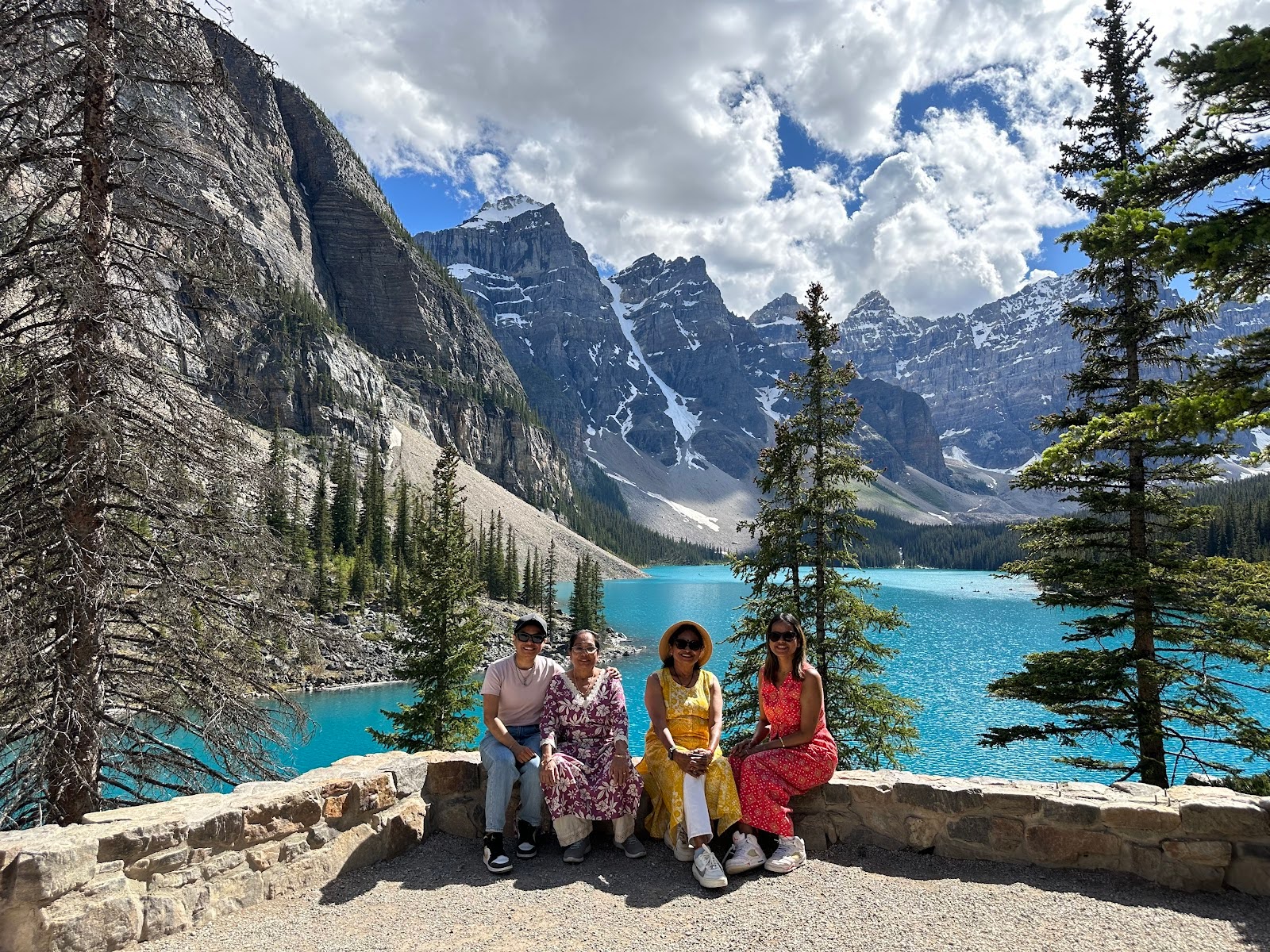 Four women sit on the rocky edge of a viewpoint overlooking a turquoise lake and mountain range.