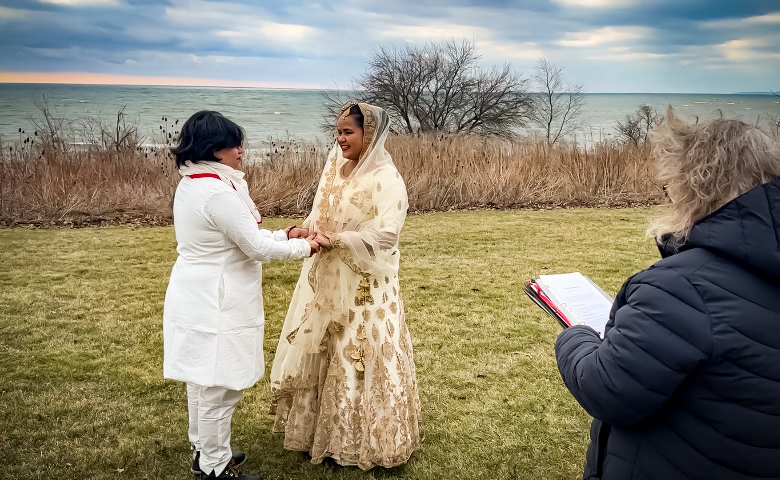Two people stand outdoors in wedding attire with water in the background. It&rsquo;s a windy day, and to the right a marriage officiant is wearing a black jacket with their back turned to the camera.