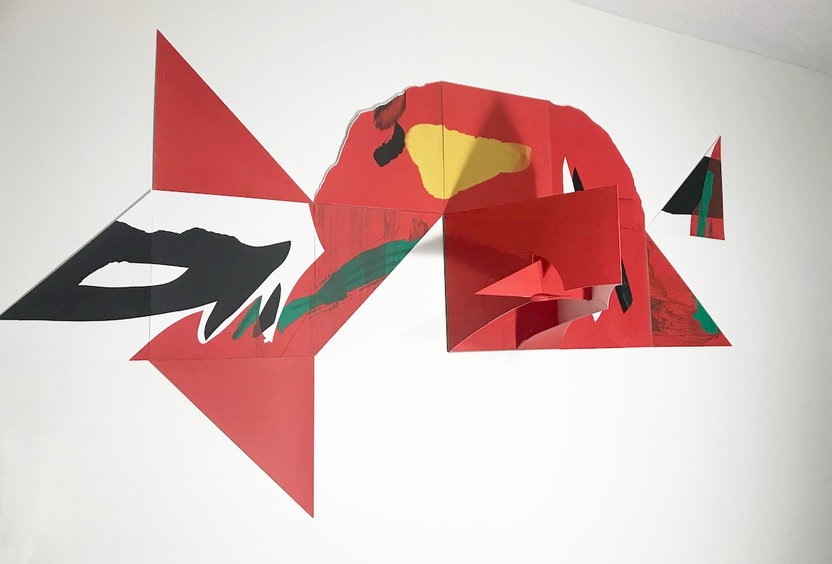 An abstract piece of artwork is mounted onto a white wall. It deploys bold red, black and green geometric shapes.