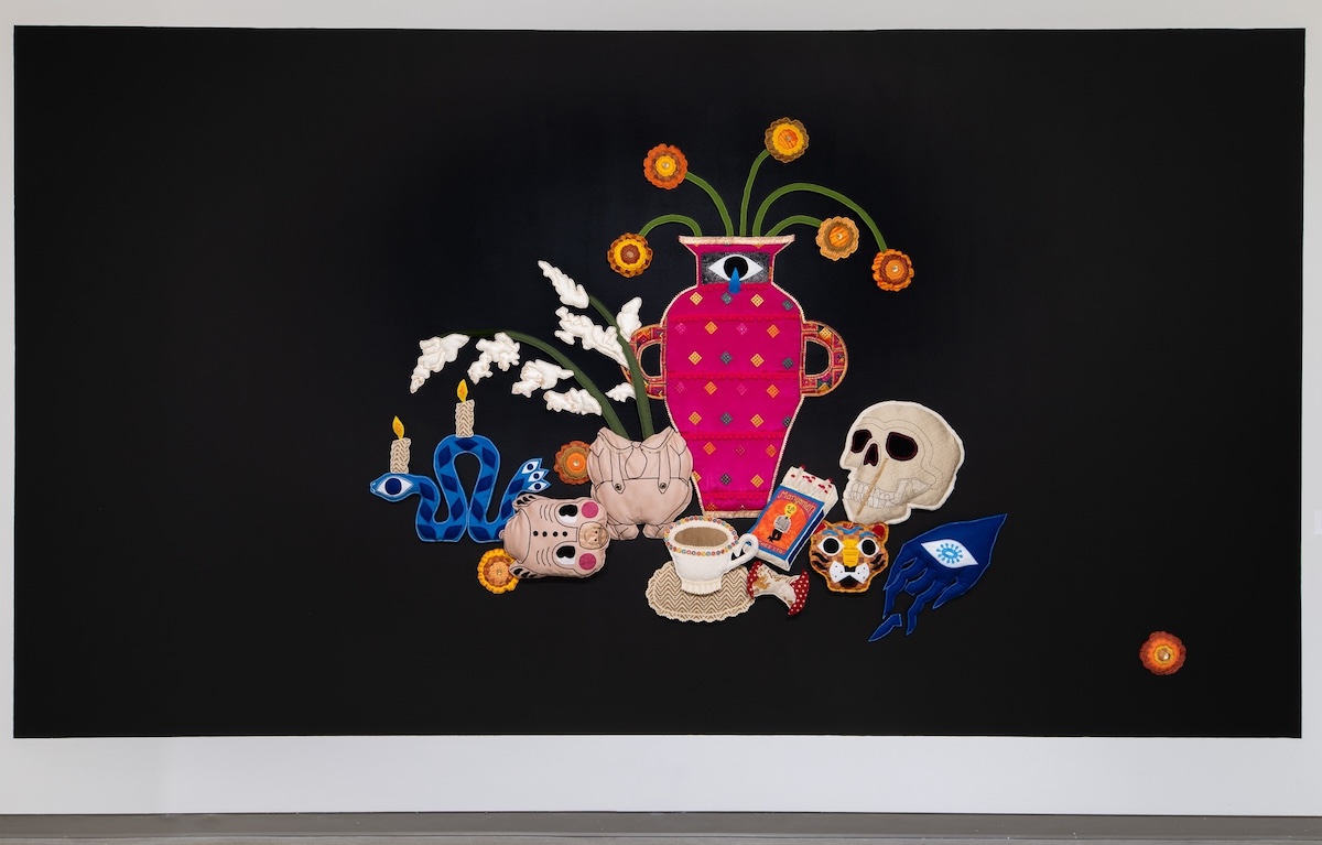 A piece of textile artwork features a fuchsia vase and other still-life objects, including a skull, a tiger's head and a cup of tea, against a wide black background. The art is hung on a white wall in a gallery space with a grey floor.