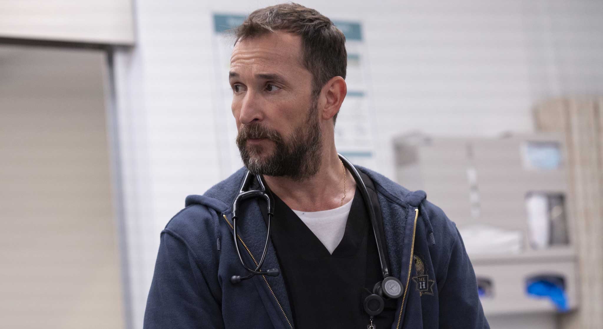 Noah Wyle has short brown hair and is wearing a navy cotton hoodie over black scrubs and a white T-shirt. A stethoscope hangs around his neck and he is looking towards the left of the frame, standing in a hospital settting.