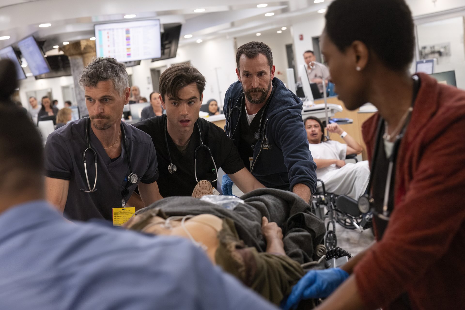 A still from The Pitt features five medical staff wheeling a person on a stretcher through an emergency department.