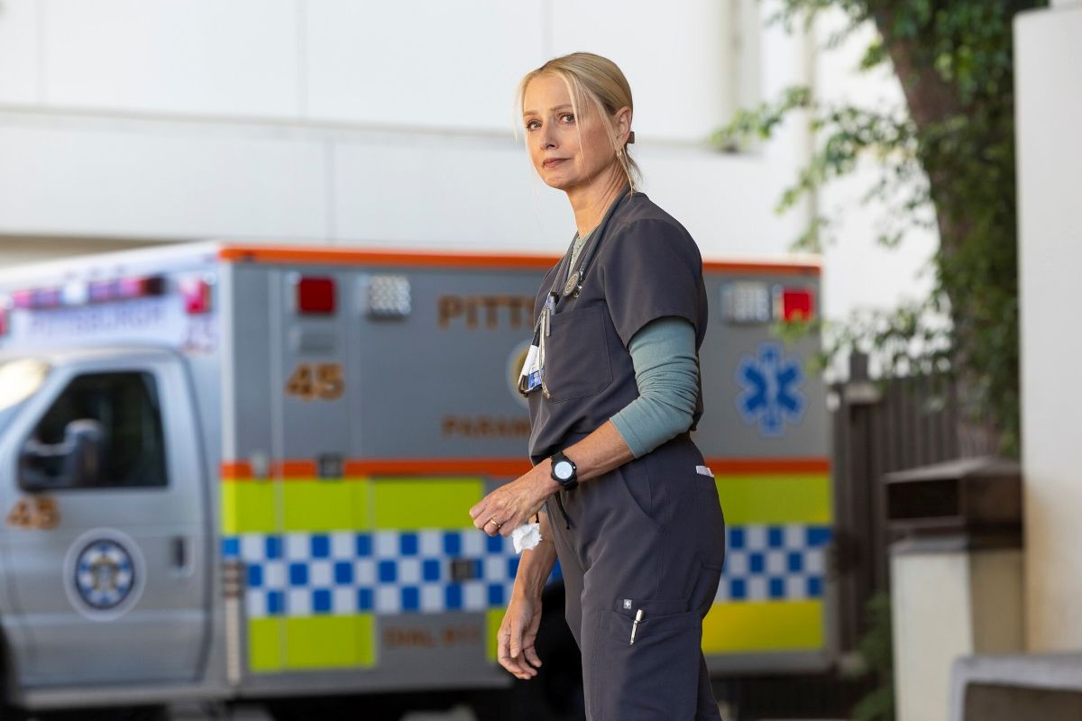 Actor Katherine LaNasa is wearing dark grey scrubs over a light turquoise long-sleeved t-shirt. She has light blond hair and is standing outdoors with an ambulance in the background.