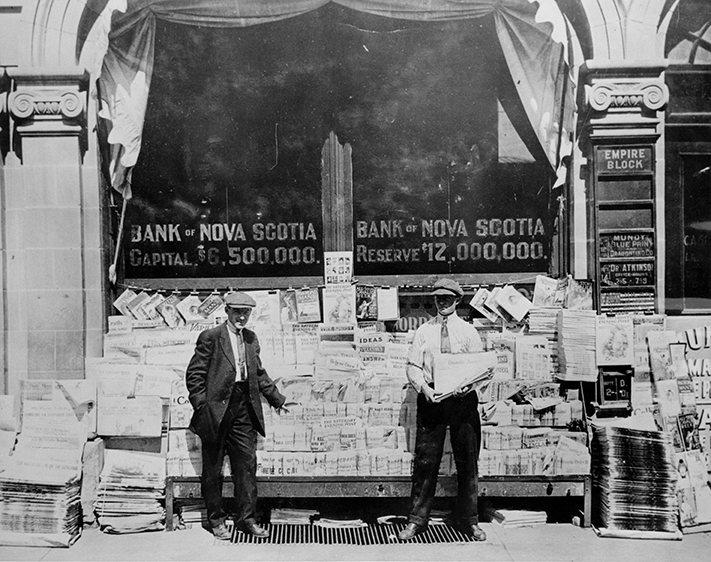 A black-and-white archival photo from 1913 features two men in pageboy caps standing in front of a news stand on an Edmonton street.