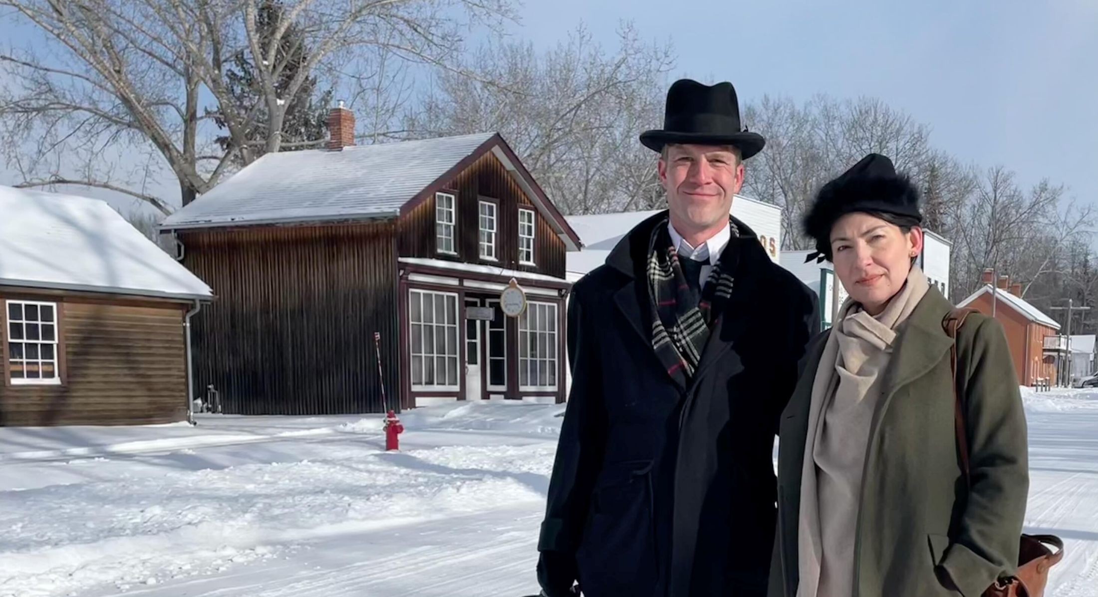 Two people stand together to the right of the frame. They are standing on a snowy street in a reconstructed town and period costumes from 1905. At right, an actor playing Balmer Watt wears a black hat and black peacoat over a white shirt; at left, an actor playing Peggy Watt wears a black hat and a long olive-green winter coat.