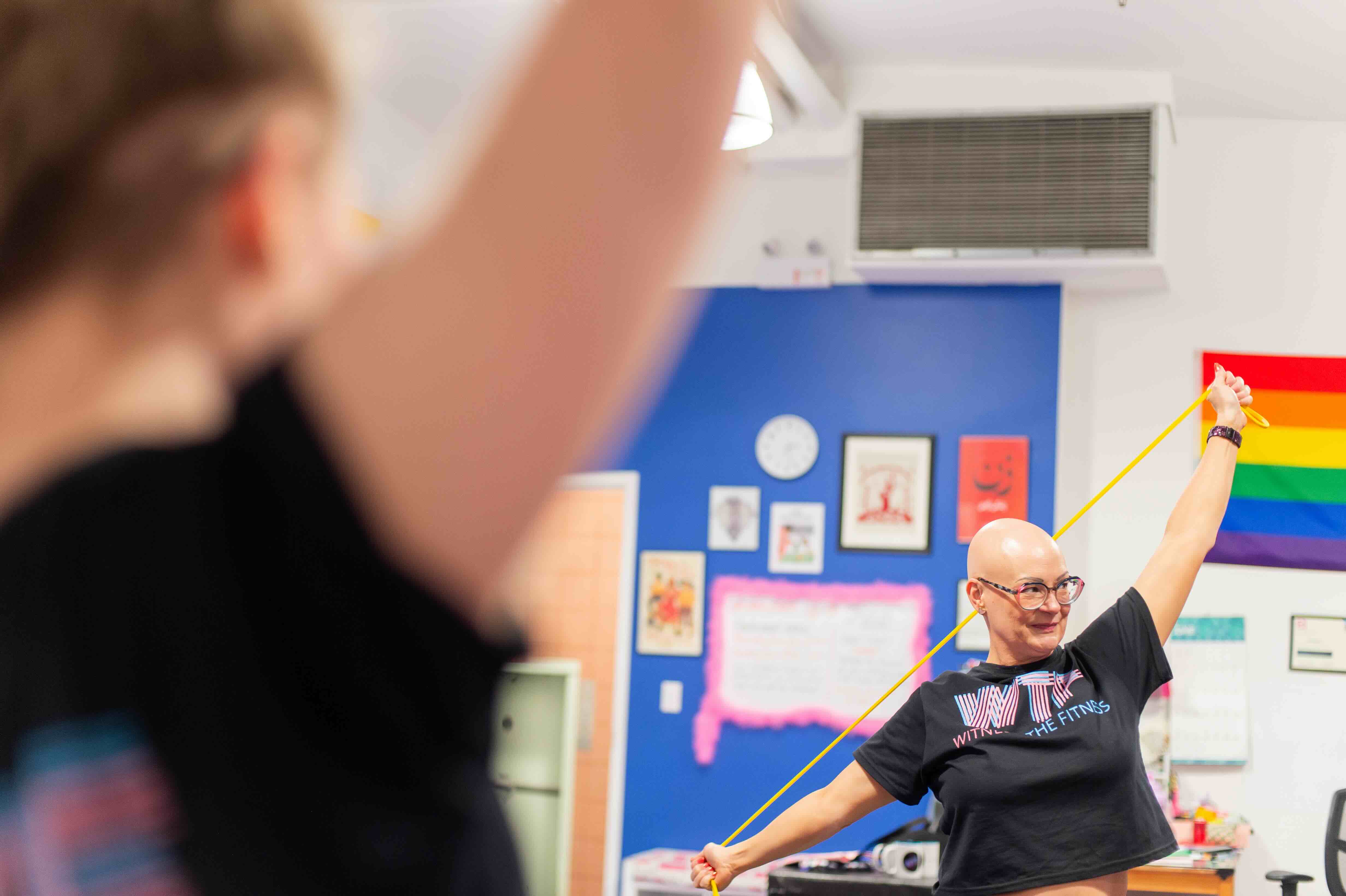 Jenn Farrell stretches a thin yellow resistance band with her arms and looks up towards the right of the frame. In the foreground a person with brown hair and a black T-shirt is mimicking a similar motion.