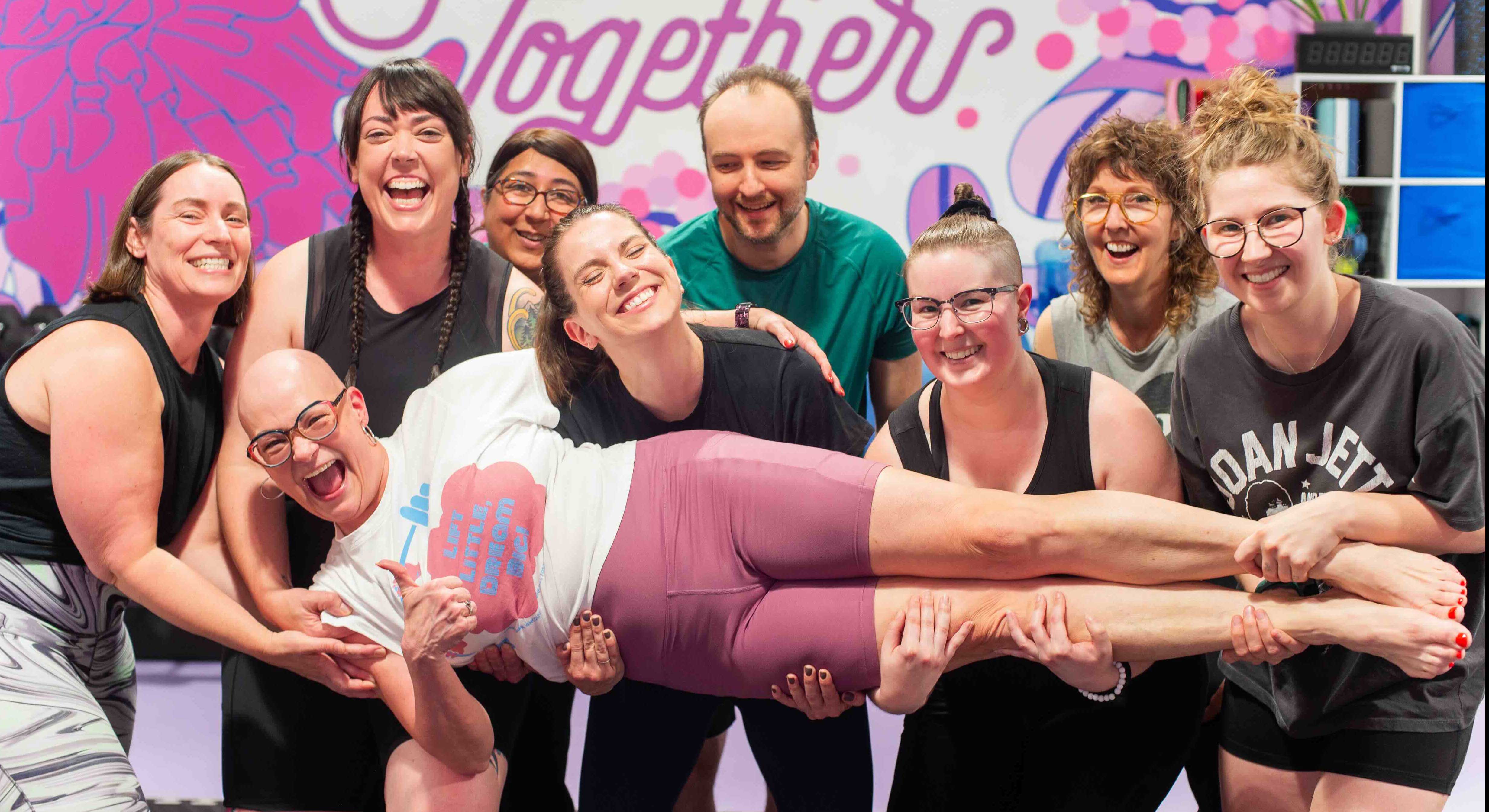 Jenn Farrell is lying horizontally in the arms of several people who are all smiling and laughing. Farrell has glasses, no hair and she is wearing a white T-shirt and lilac bicycle shorts.