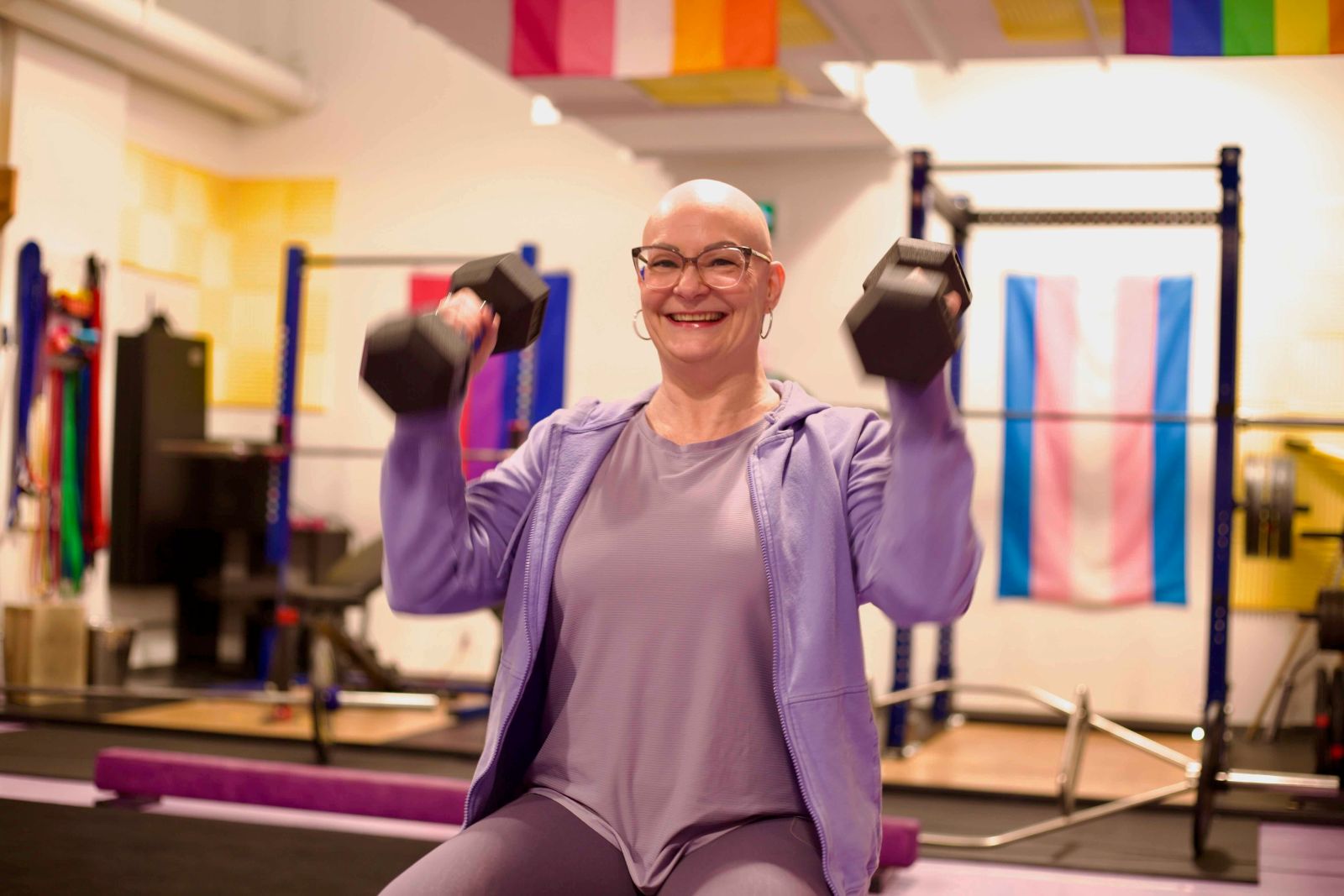 Jenn Farrell is wearing a lilac hoodie over a matching T-shirt and leggings. She is seated on a bench in a gym lifting two black weights. She is smiling at the camera.