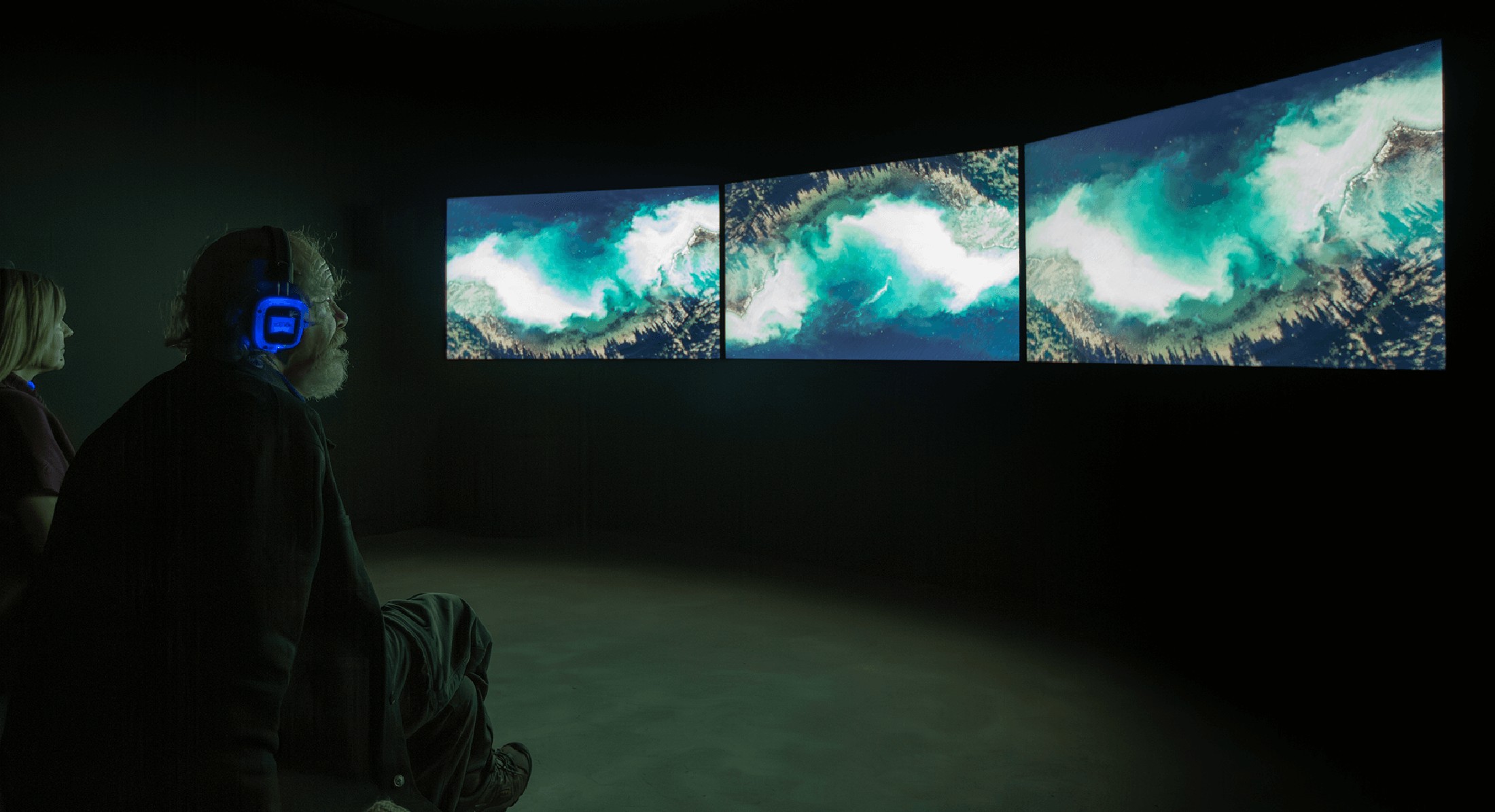Two people wearing headphones sit on benches watching three screens that show water flowing, with herring spawn, near the shore. 
