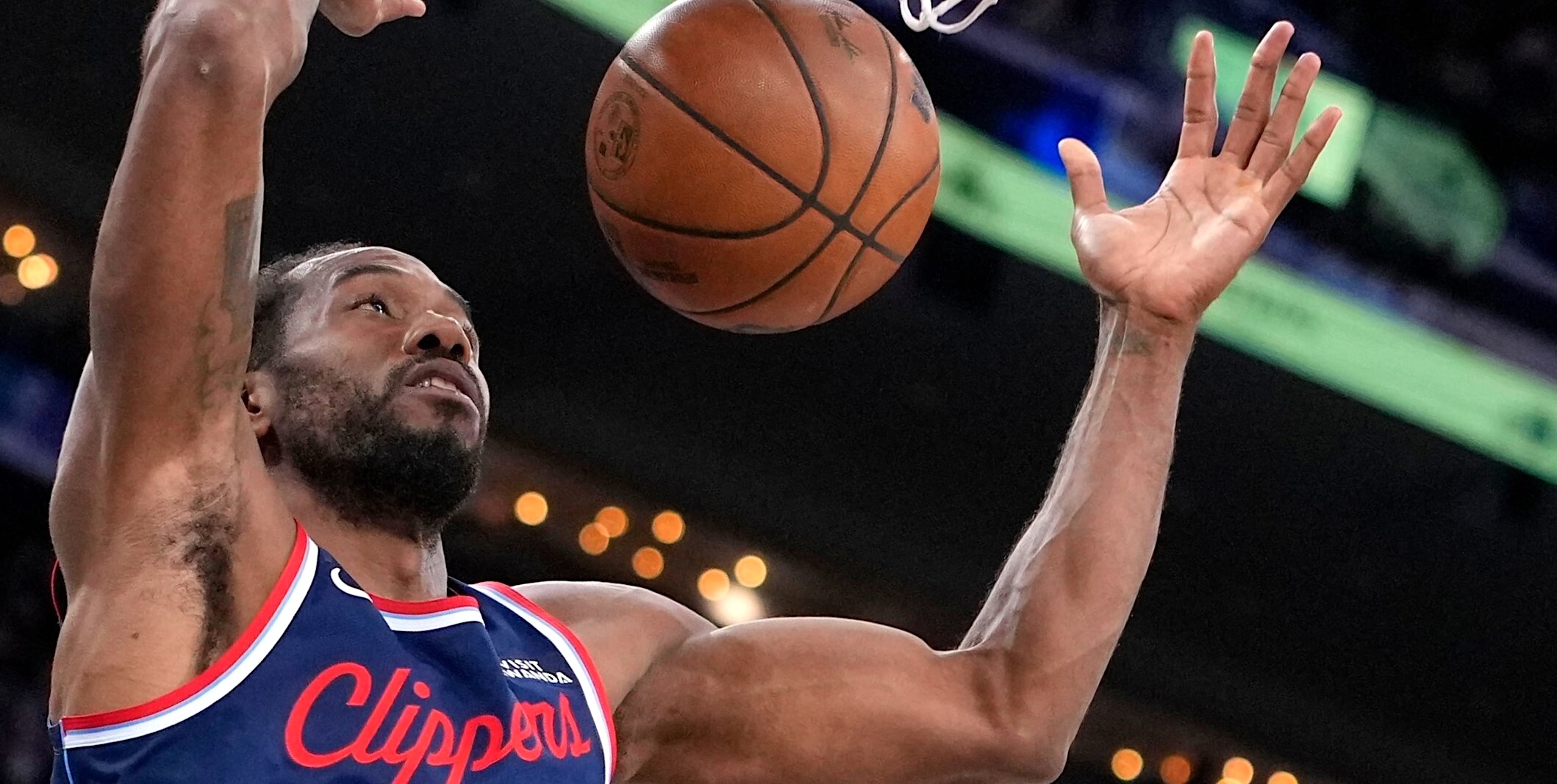 A close-up press photo of Los Angeles Clippers forward Kawhi Leonard depicts the athlete in mid-air as he dunks a basketball through the net. Leonard has dark hair and a beard and he is jumping while raising his arms around the net.