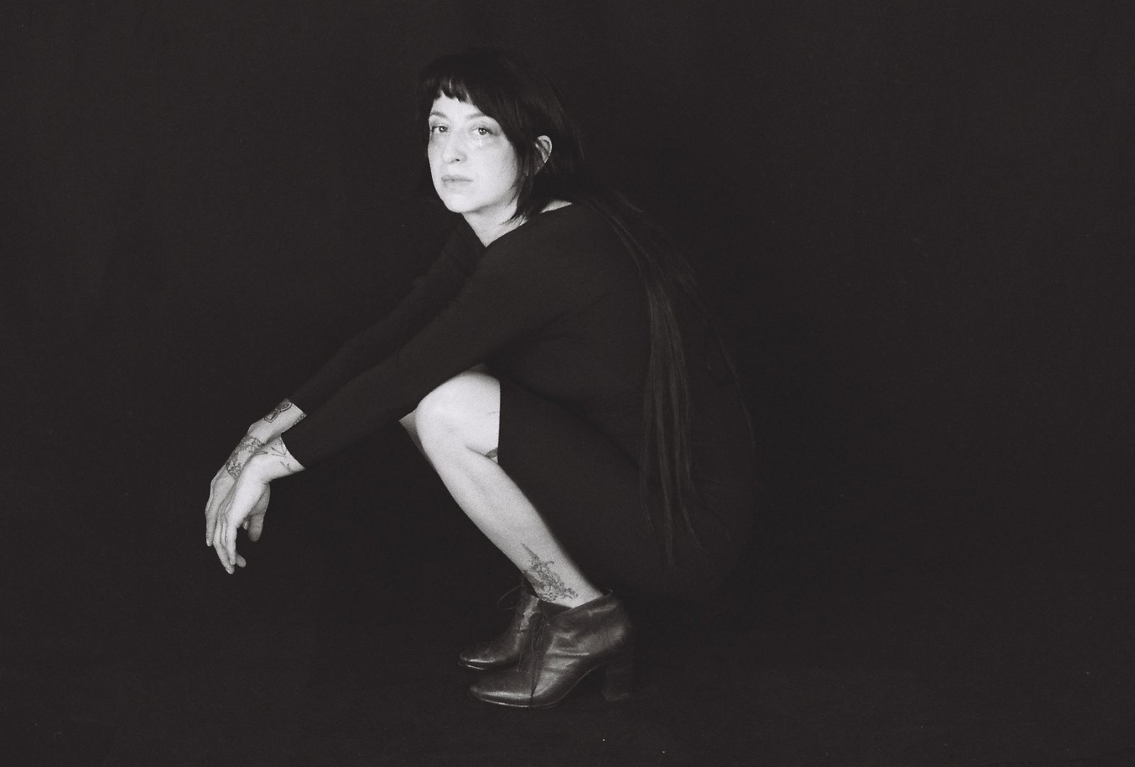 A black-and-white studio portrait of Ora Cogan features Cogan posing against a black studio background. She is crouching with her arms over her knees facing the right of the frame. She has long dark hair and she is wearing a black dress and ankle boots.