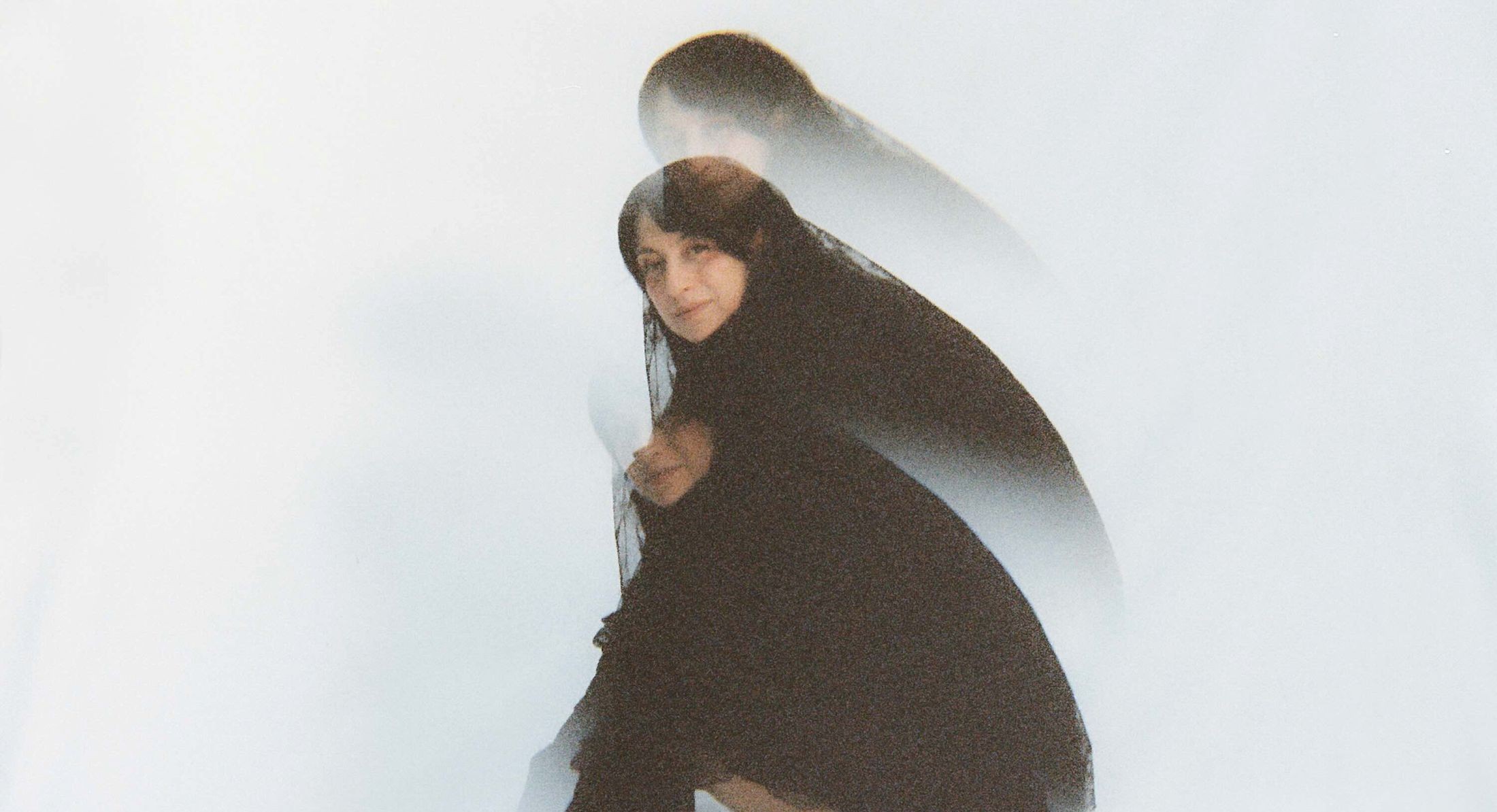 An abstract colour photograph features three stacked, overlapping images of Ora Cogan against a white background. She is crouching, hands towards the floor, and facing the right of the frame. She turns to face the camera and she is smiling. She has long dark hair and is wearing a black dress and ankle boots.