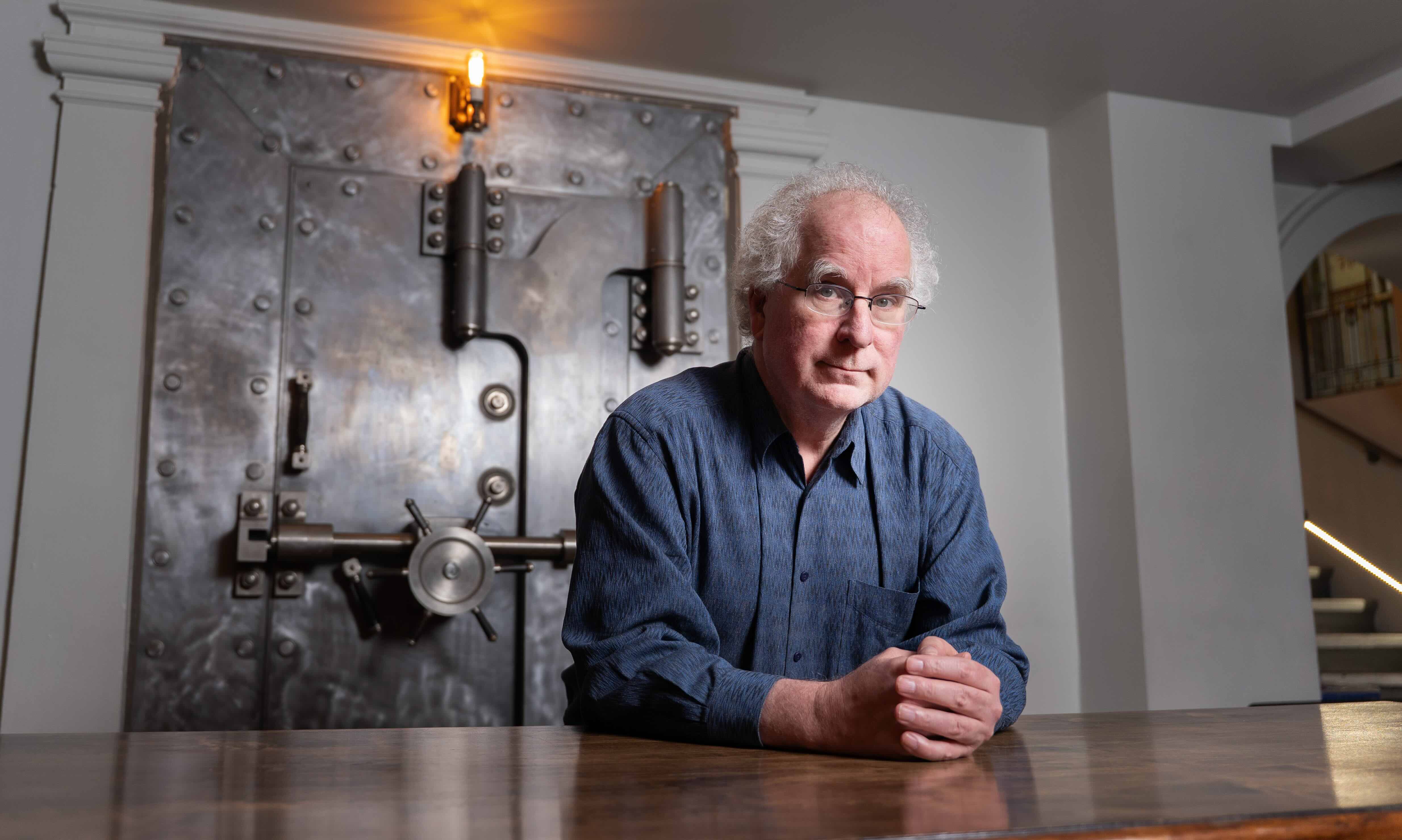 Brewster Kahle is seated at a wooden table. He has curly white hair, glasses and a blue button-down shirt. Behind him is the metal door to a large vault, above which shines an antiquated-looking orange light.