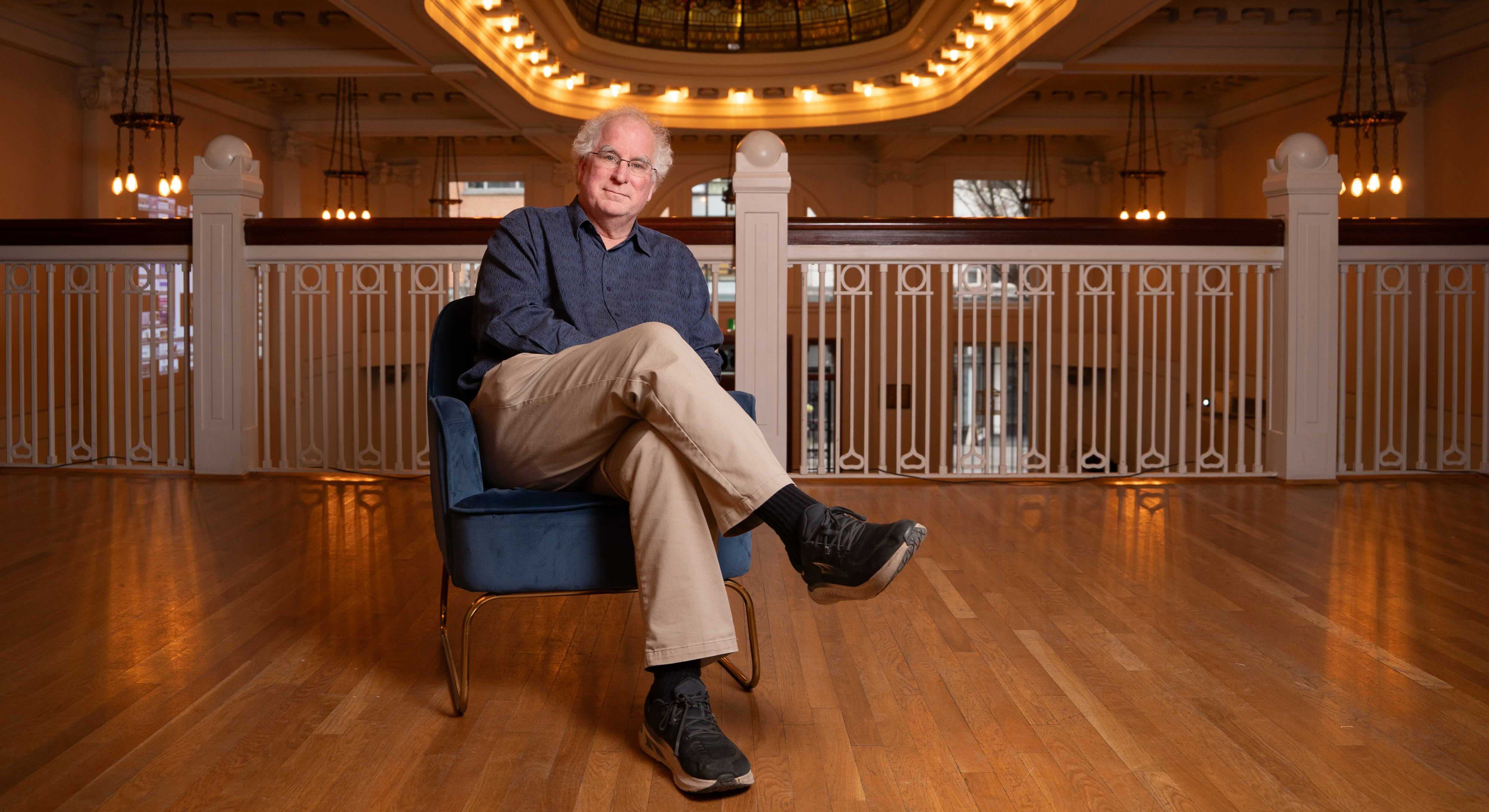 Brewster Kahle is seated on a low blue chair with his legs crossed. He has short white curly hair and glasses, and he is wearing a blue button-down shirt and khaki trousers. Behind him is a white balcony railing and above him is an orate ovular light fixture. 