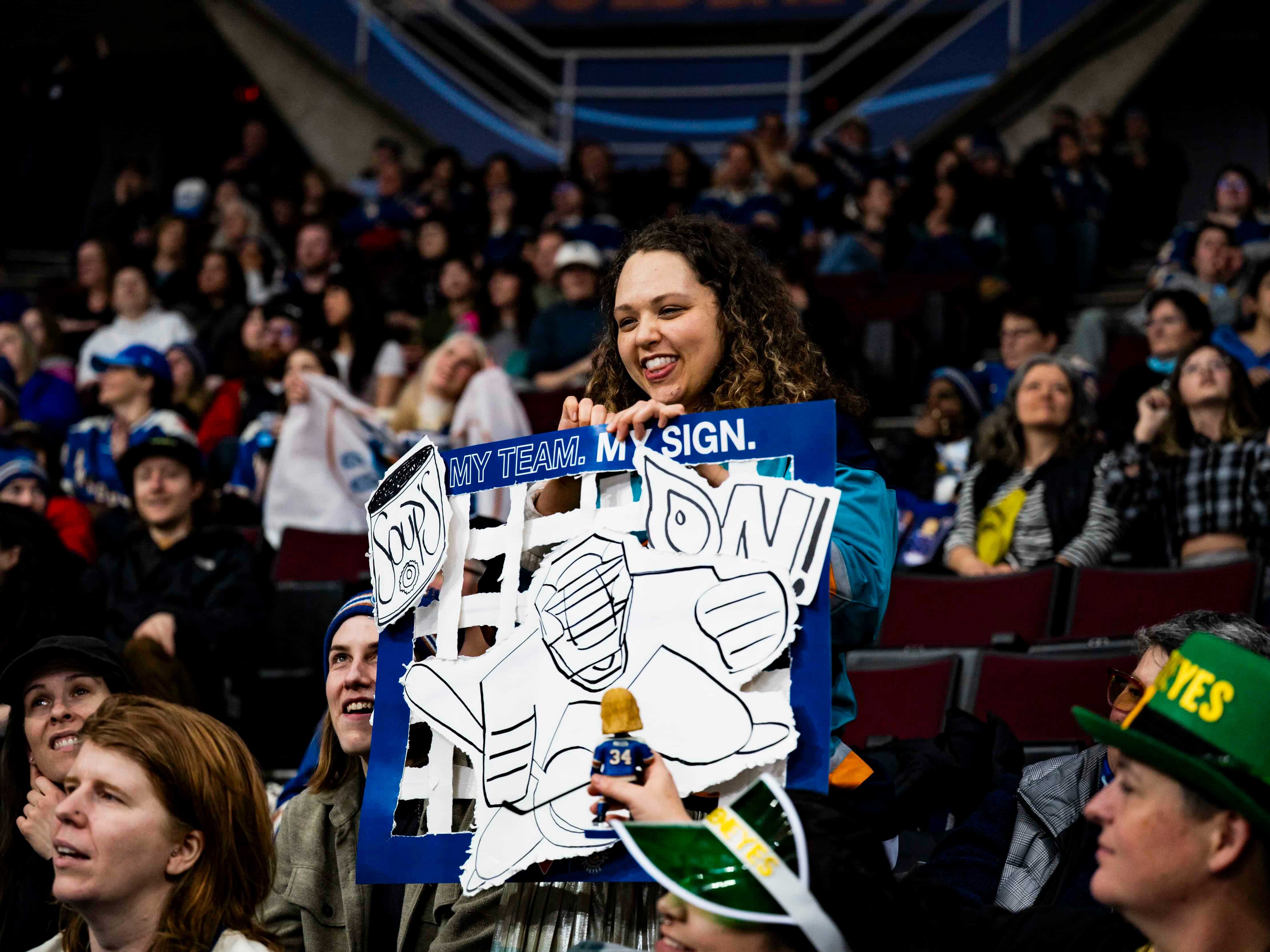 A woman with black curly long hair holds up a sign at a hockey stadium. The sign reads &ldquo;soup&rsquo;s on!&rdquo; and includes a hand-drawn image of a goalie.